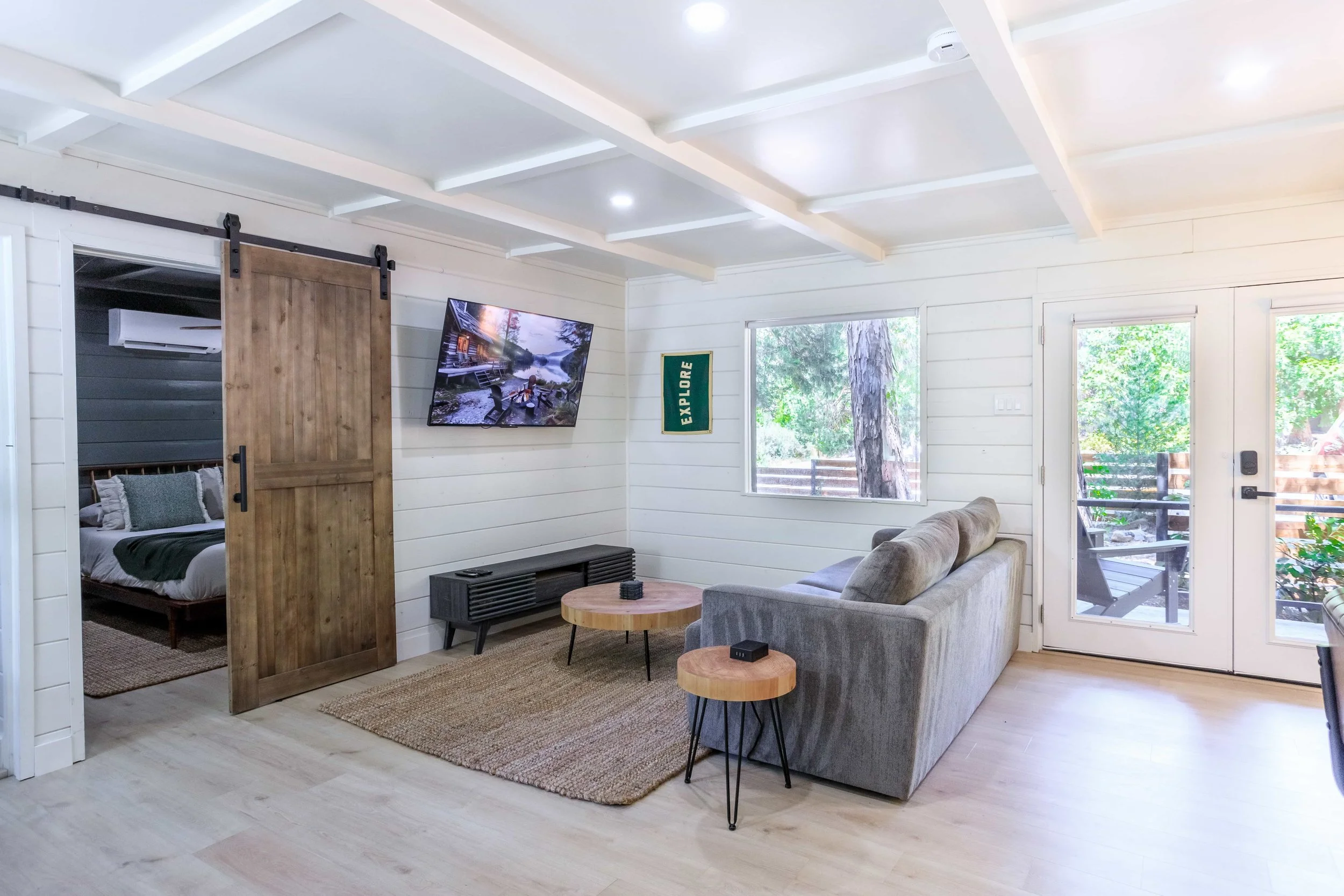 Living room with a gray sofa, two small round wooden tables, a wall-mounted TV, a window with a view of trees, and a sliding barn door leading to a bedroom.