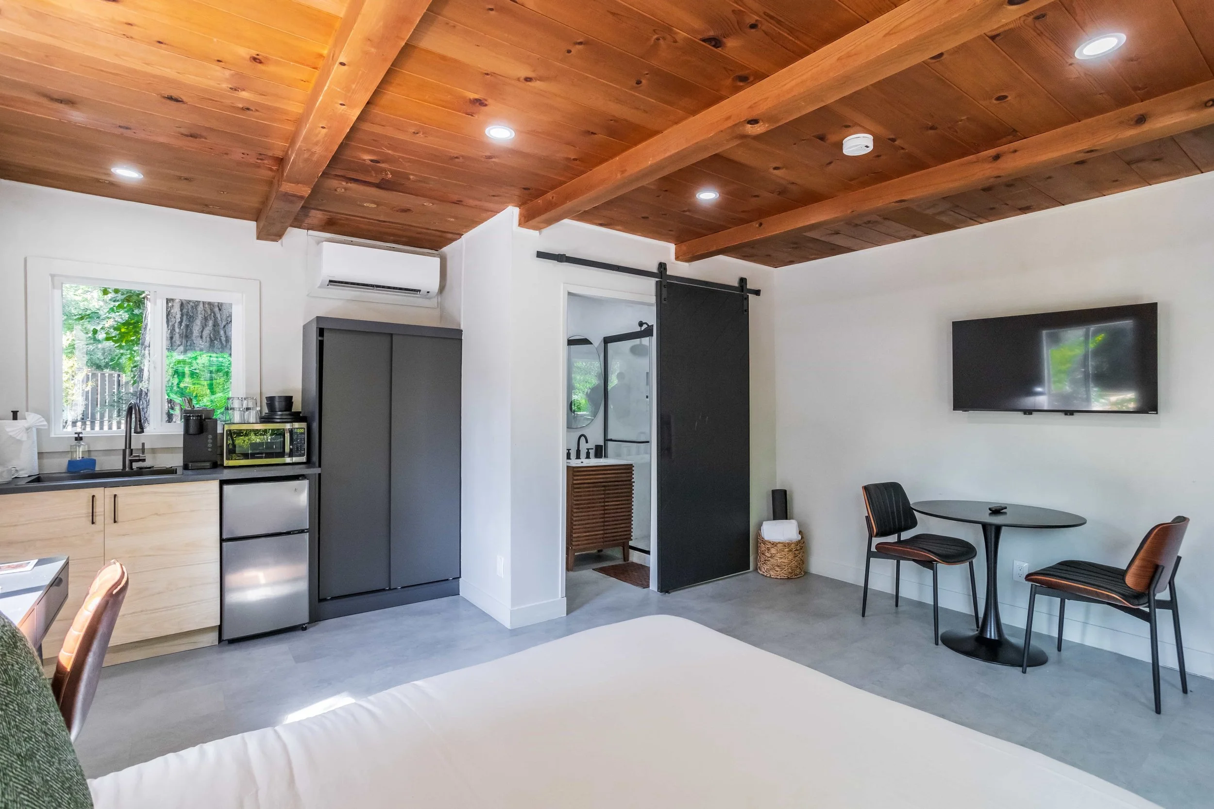Interior of a modern studio apartment featuring a kitchenette with a window, a small dining area with two chairs and a round table, a wall-mounted flat-screen TV, and a wooden ceiling with recessed lighting.