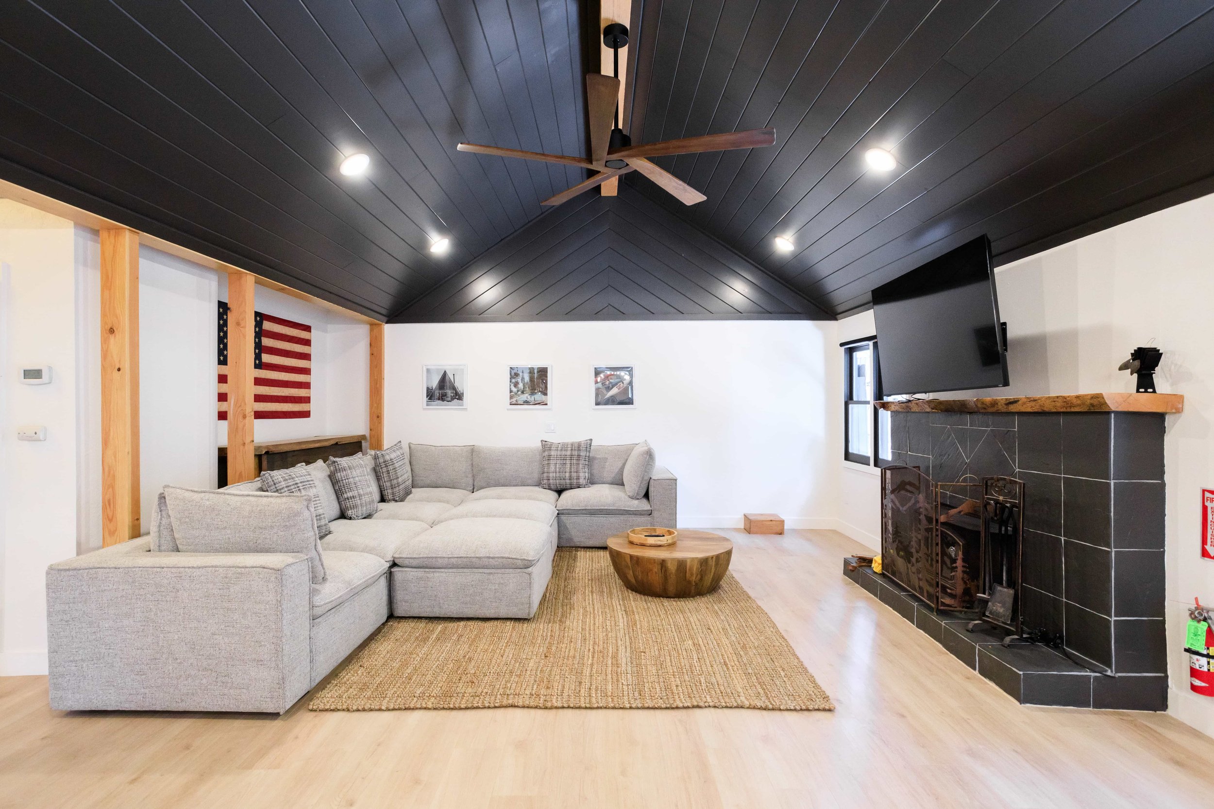 The Pinetree Hotel Idyllwild: a - Idyllwild California, cozy cabin, mountain resort Living room featuring a sectional sofa, wooden coffee table, black tiled fireplace with a wooden mantel, wall-mounted TV, and a dark ceiling with a ceiling fan.