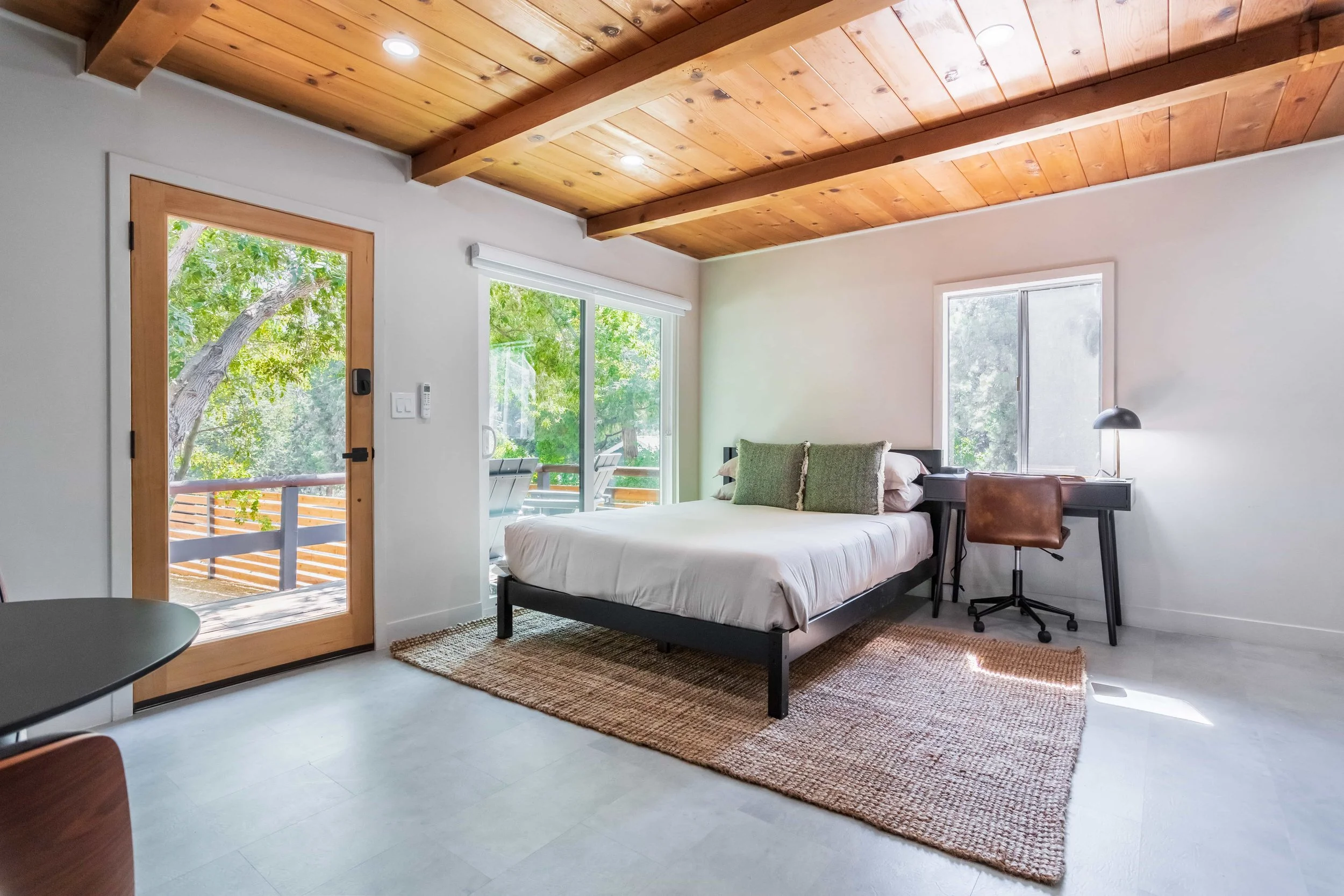 Bedroom with white walls, wooden ceiling, and large windows showing green trees outside, containing a bed with green pillows, a side desk with a lamp, a brown chair, and a glass door leading to a balcony.