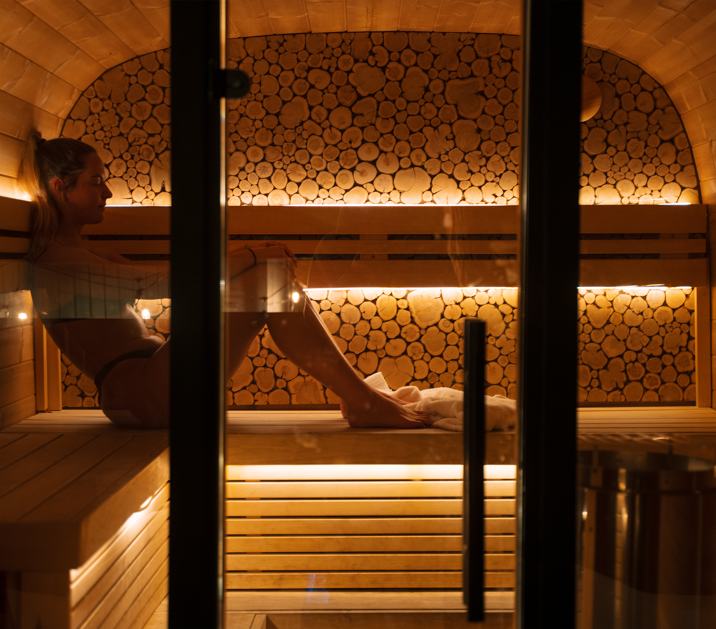 The Pinetree hotel wellness: custom wooden sauna room, with a wall of logs in the background, heated lighting, and a glass door in front.
