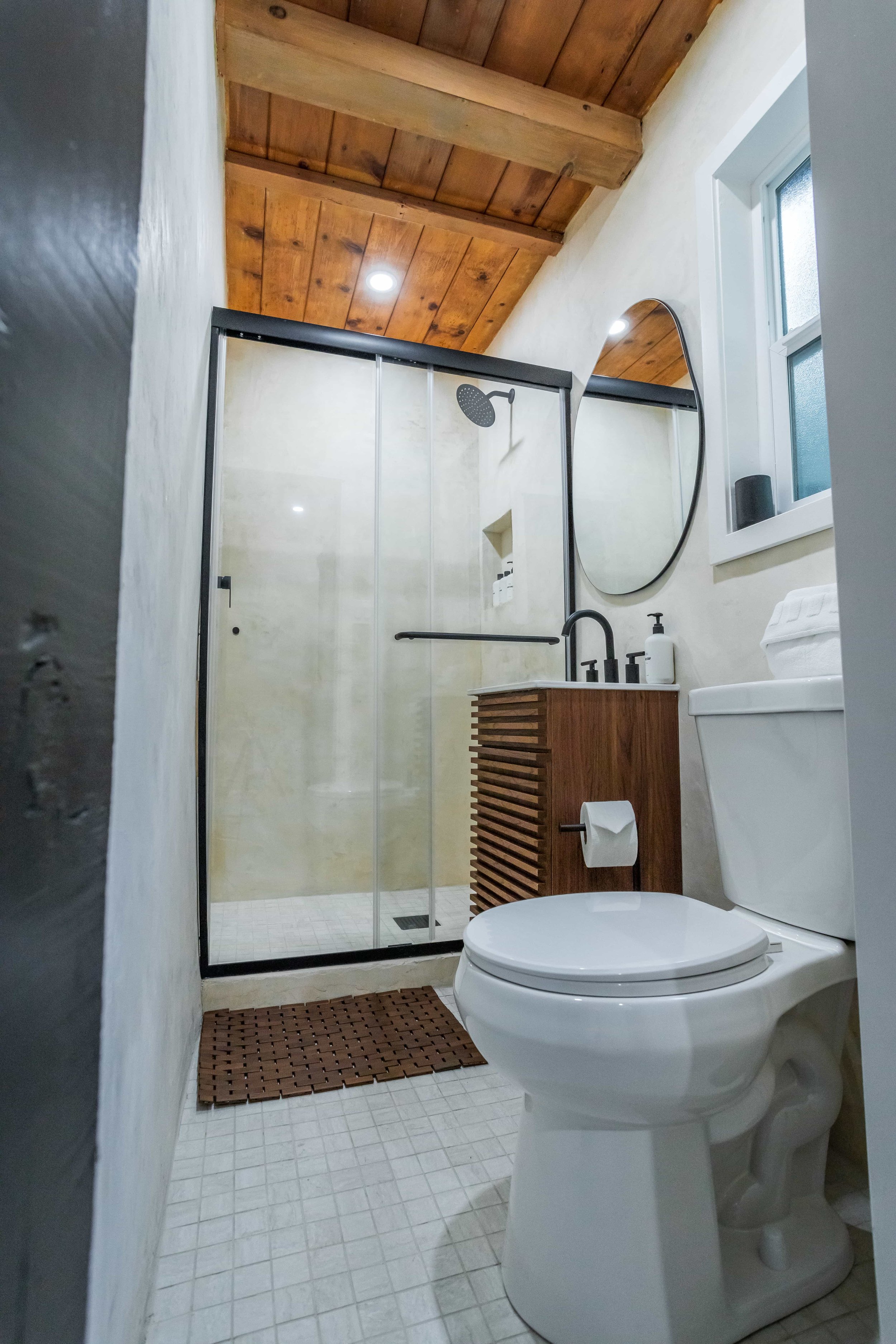 A modern bathroom with a toilet, a wooden vanity with a black faucet, a shower with glass doors, and a round mirror on the wall. The ceiling has wooden panels, and there is a window next to the mirror.