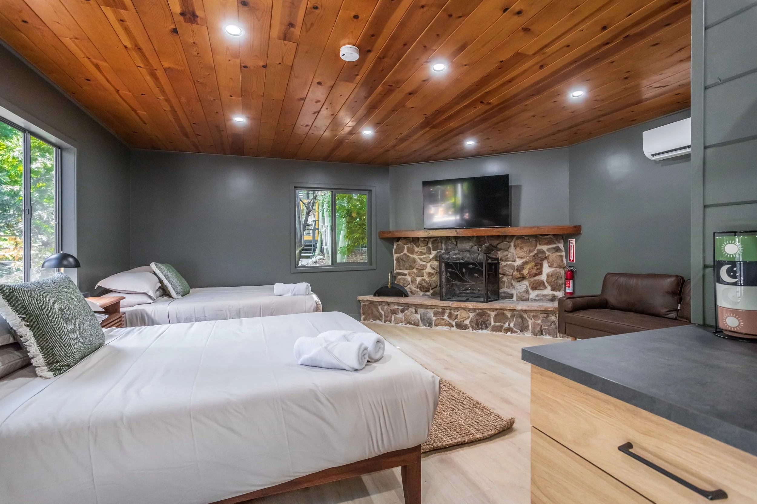 Two beds with white linens and rolled towels, a stone fireplace with a mounted TV above, and a brown leather couch in a cozy room with a wooden ceiling and gray walls.