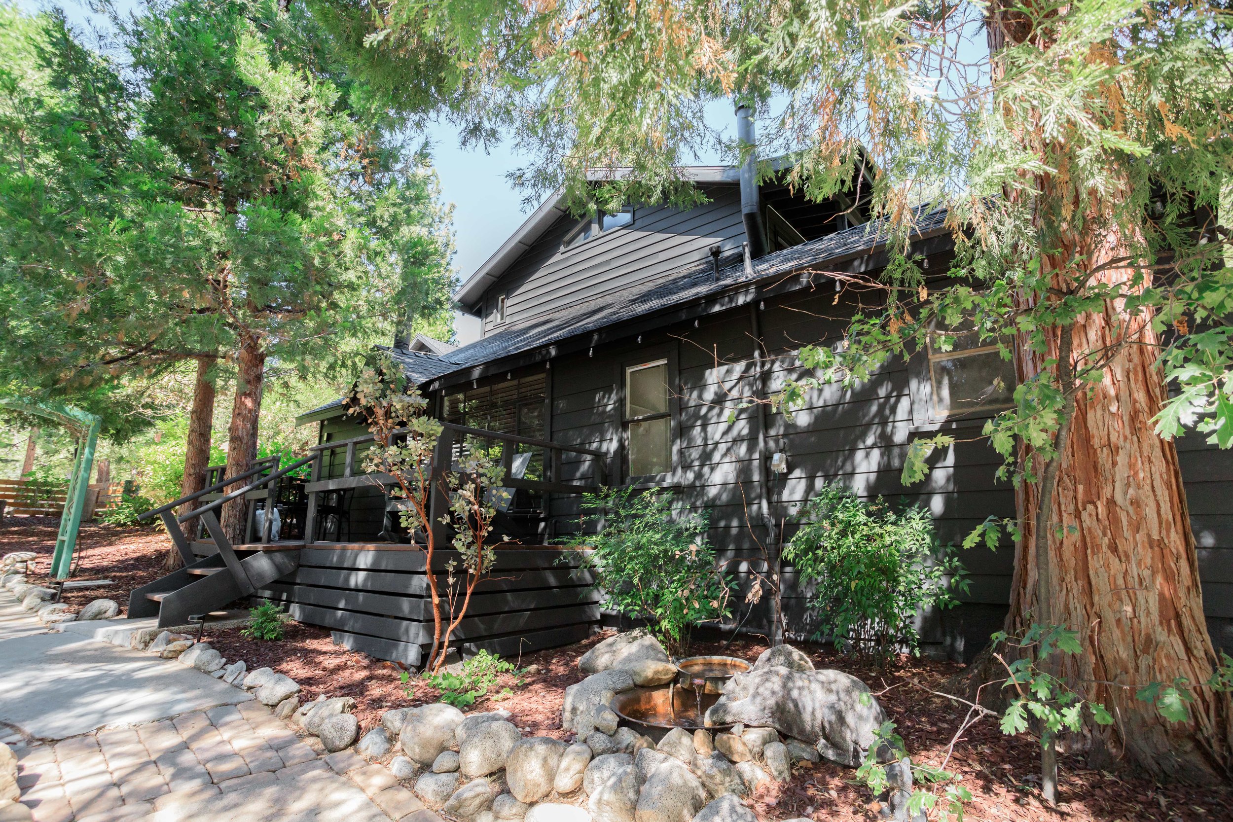 Rear view of a black house surrounded by large trees and a landscaped yard with rocks, small plants, and a patio with steps.