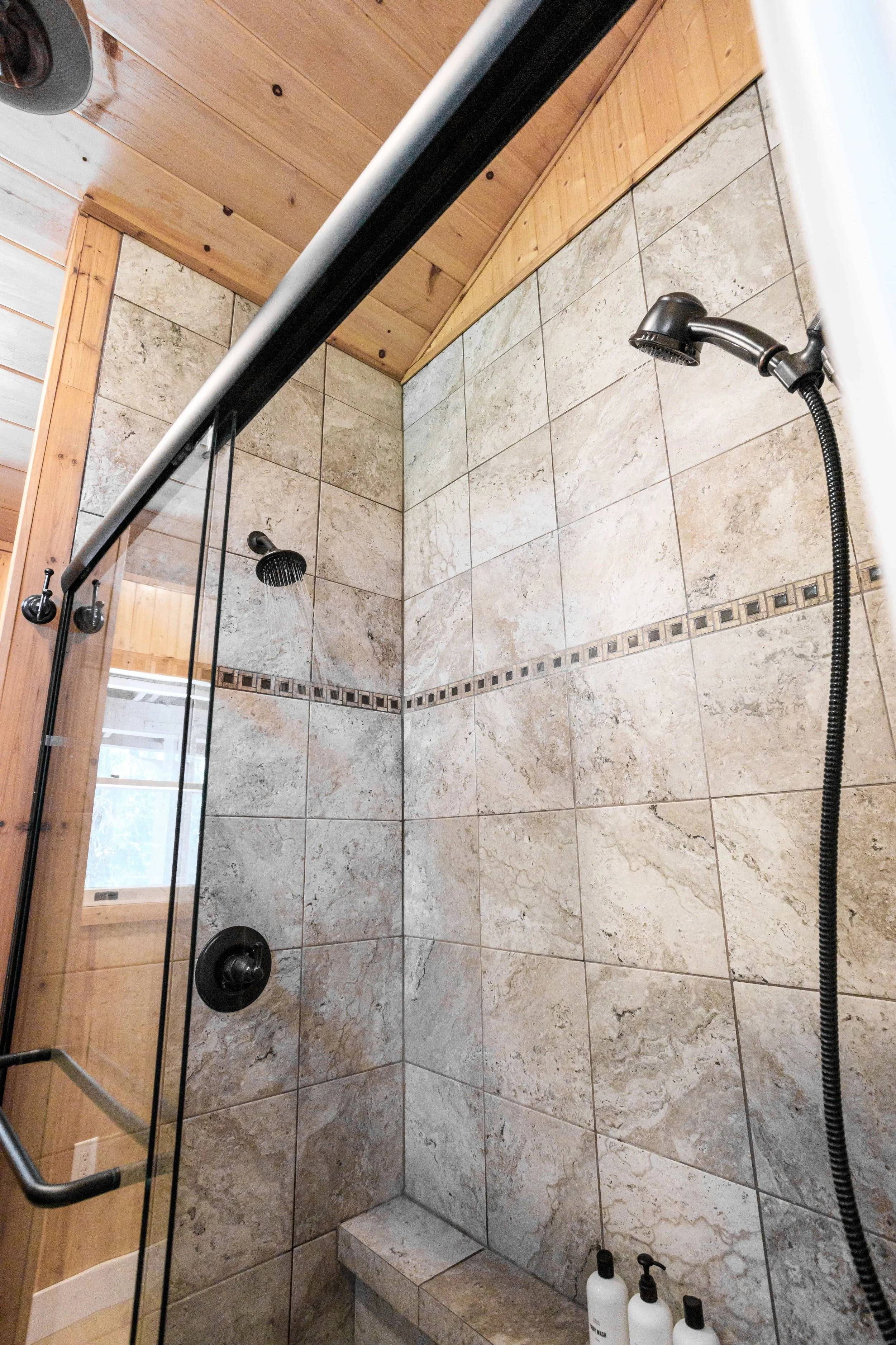 Inside a tiled shower with two showerheads, one fixed and one handheld, and three bottles of toiletries on a built-in shelf.