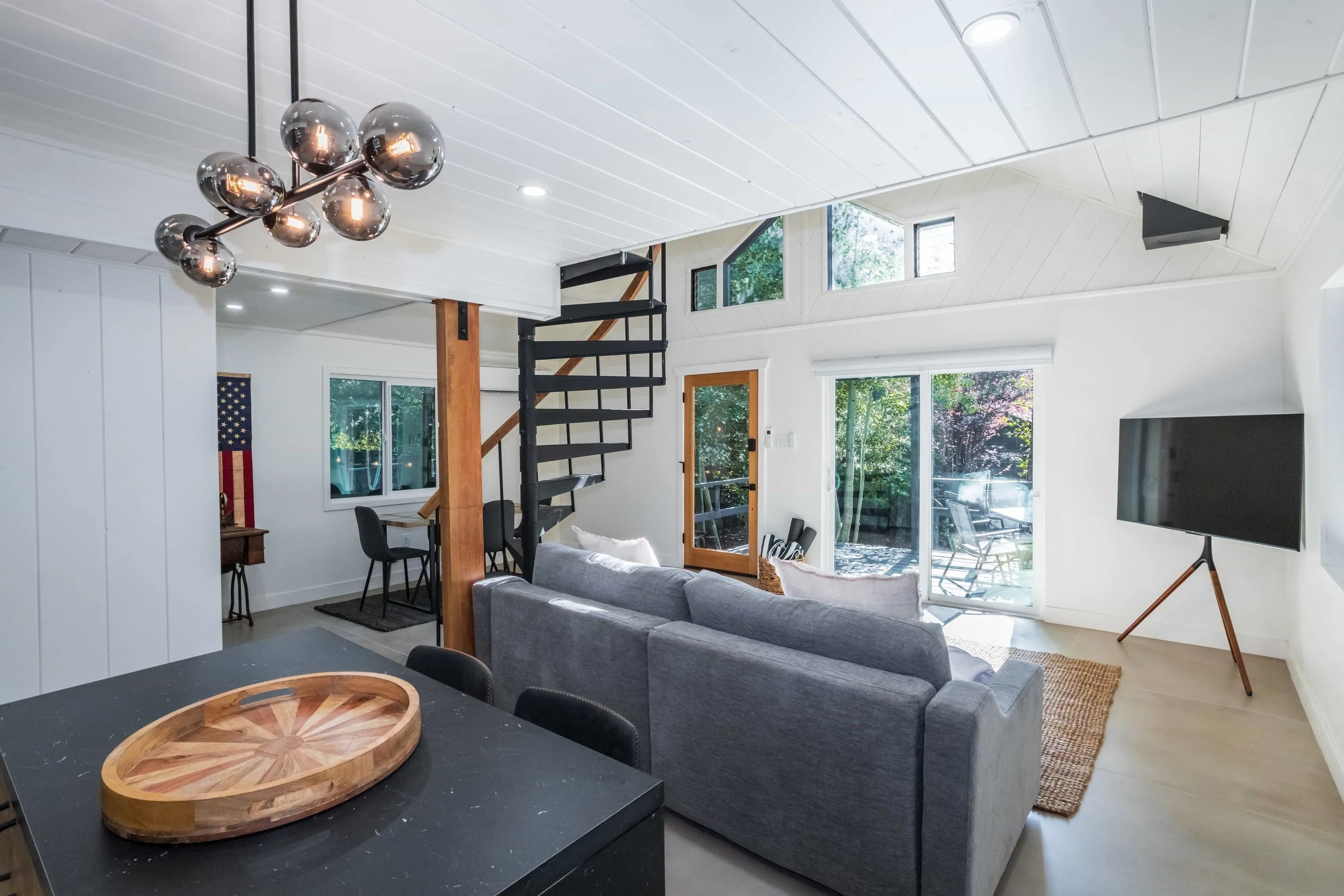Modern living room with gray sofa, TV on stand, and sliding glass doors leading outside, with a spiral staircase and dining area in the background.