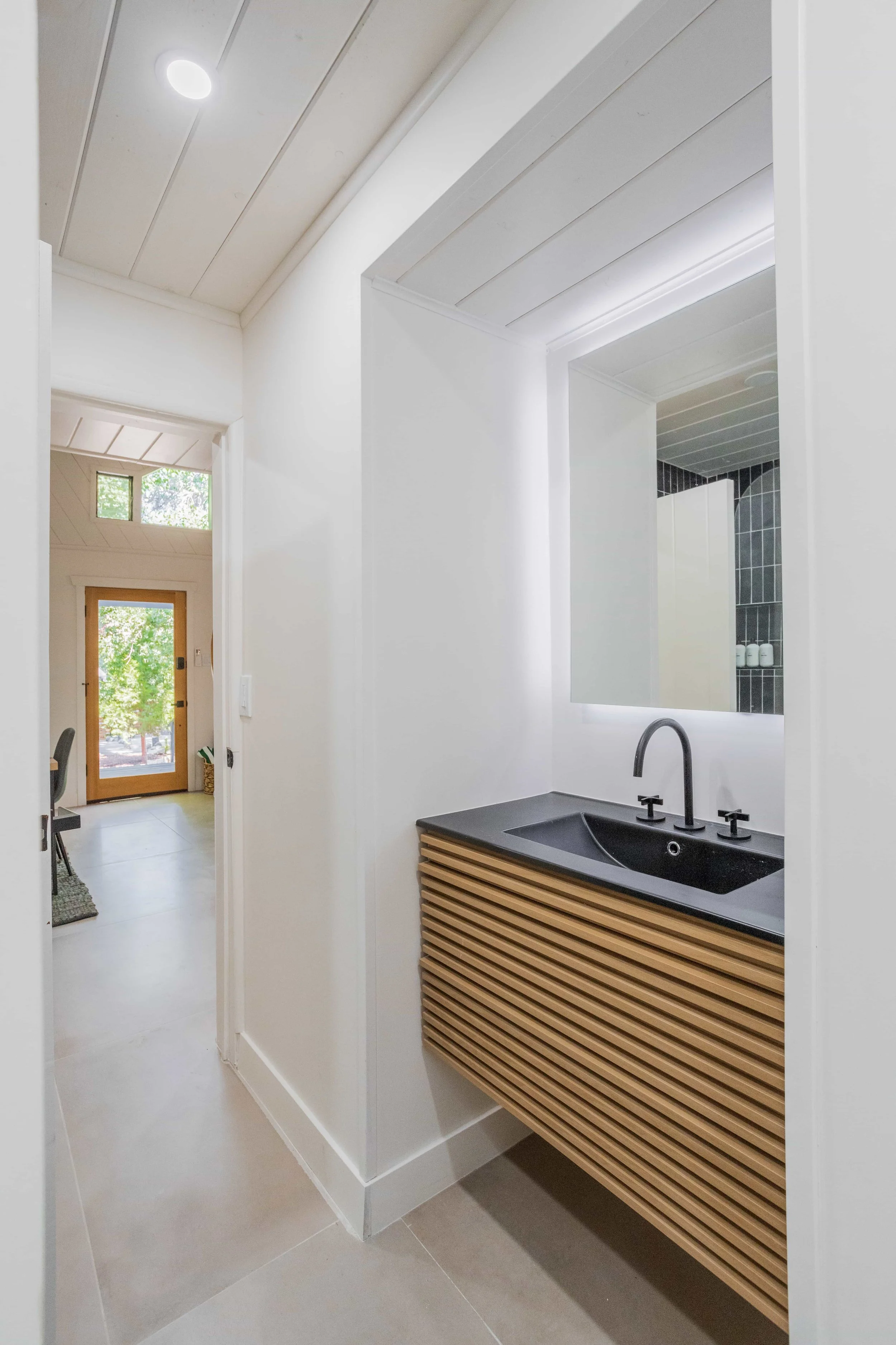Modern bathroom with black sink, mirrored backlit mirror, and wooden cabinet, with view into entryway and living space with dining table.