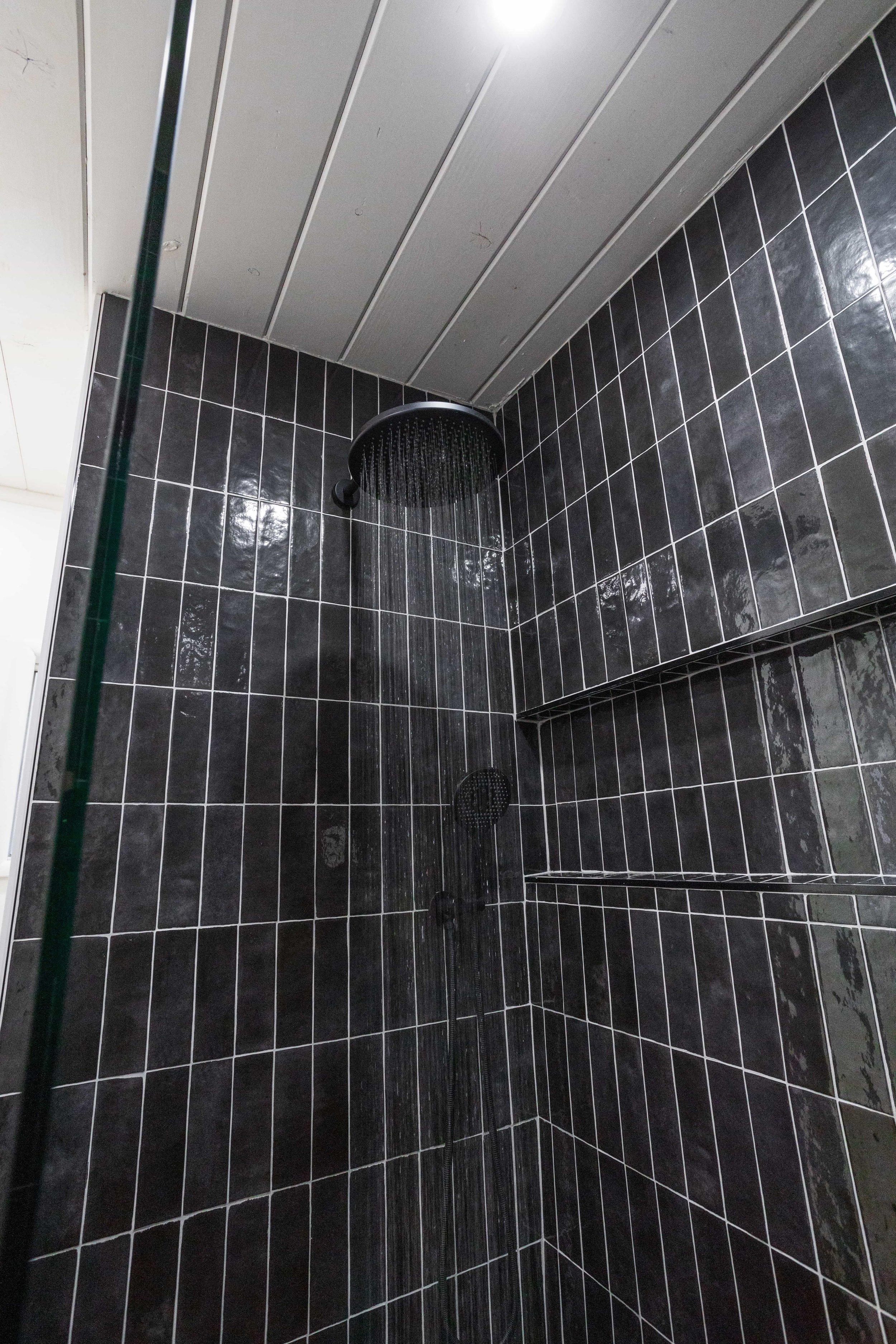 A black tiled shower with a black rain showerhead and a handheld showerhead on the wall, with a small built-in shelf in the corner.