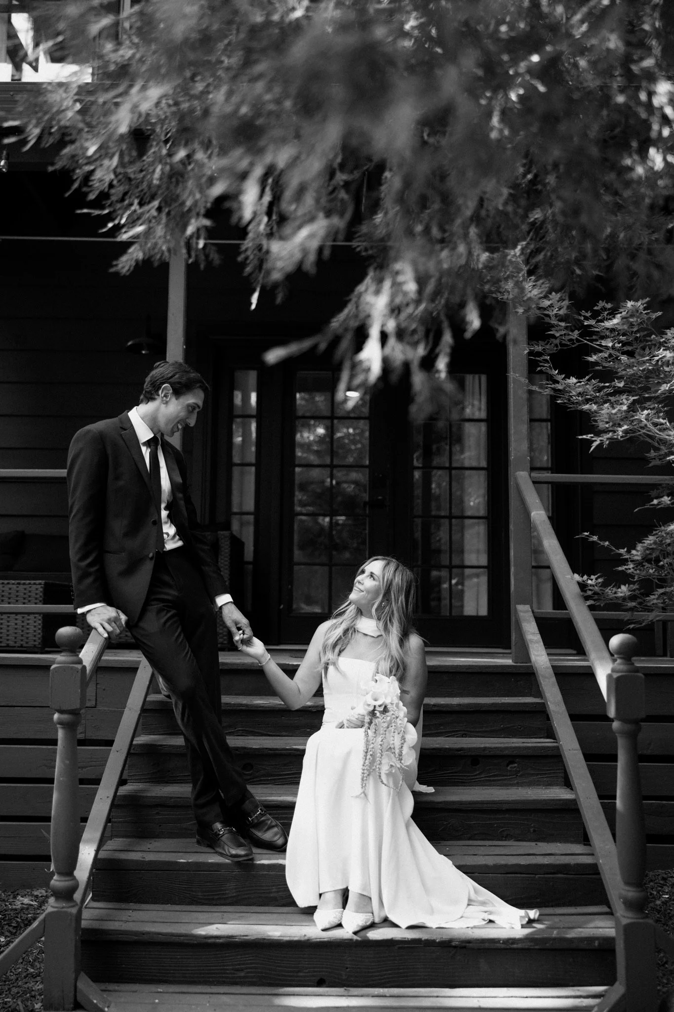 A black and white photo of a couple on a front porch staircase, with the woman sitting in a wedding dress holding a bouquet, gazing up at the man standing on the stairs, holding her hand. Trees and a house with glass double doors are in the background.