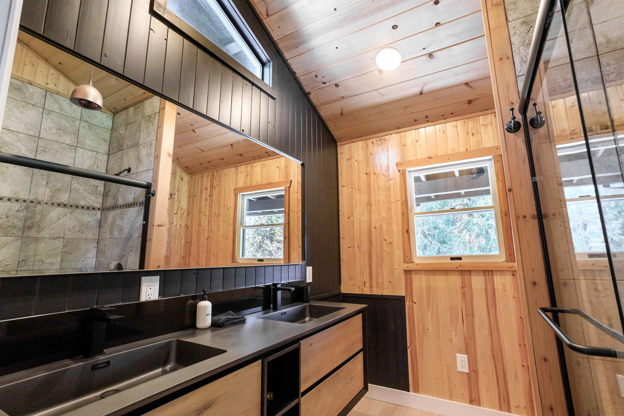 Bathroom with wood-paneled walls, a large mirror, two sinks, and two windows showing trees outside.