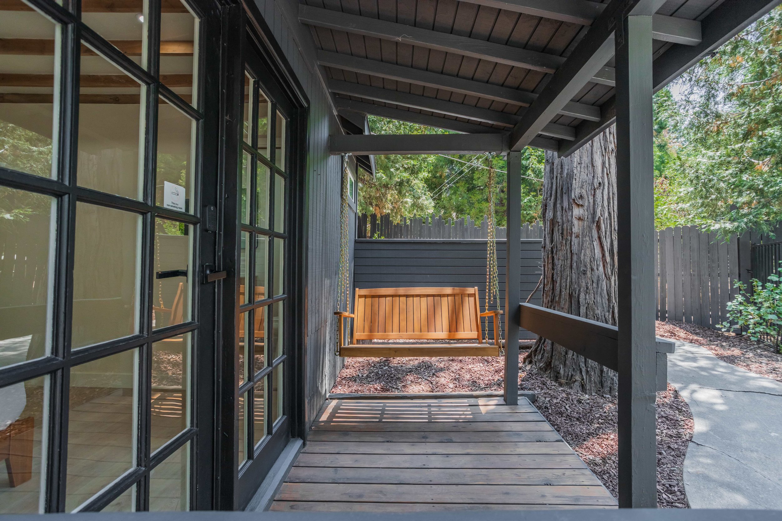 A wooden porch swing hanging from chains on a porch with wood flooring and a black exterior wall, next to a large tree and a wooden fence, with a curved concrete pathway nearby.