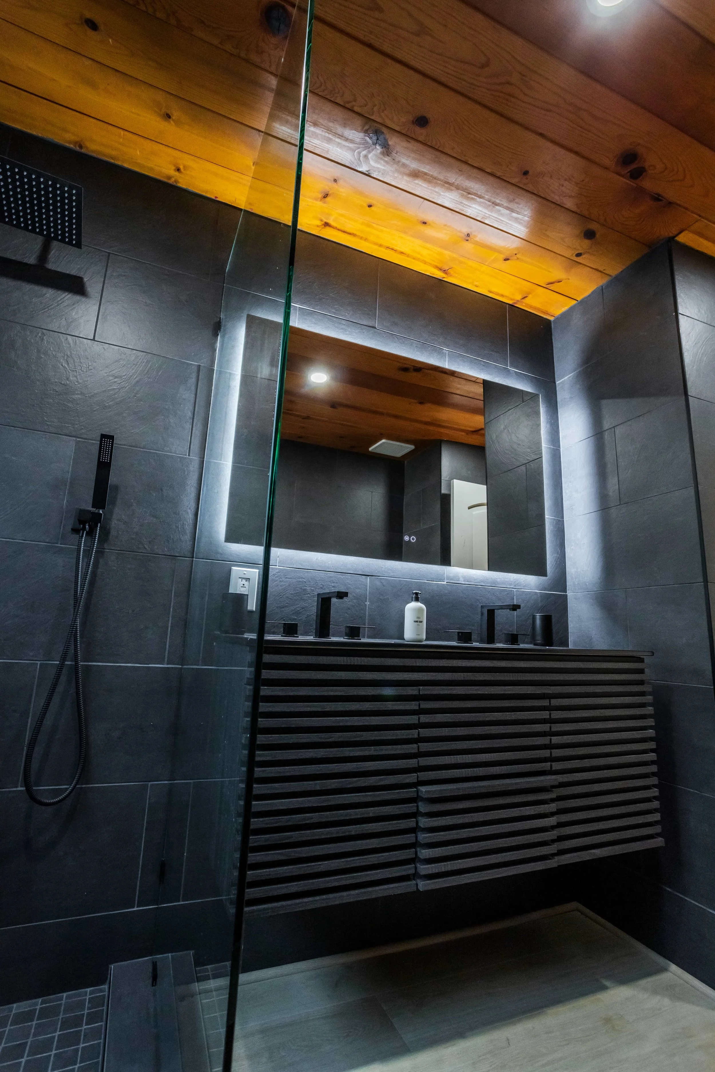 The Pinetree Hotel Idyllwild:  a - Idyllwild California, cozy cabin, mountain resort, mountain getaway Modern bathroom in cabin 1 with dark tiled walls, wooden ceiling, and walk-in shower with glass door and frosted window.