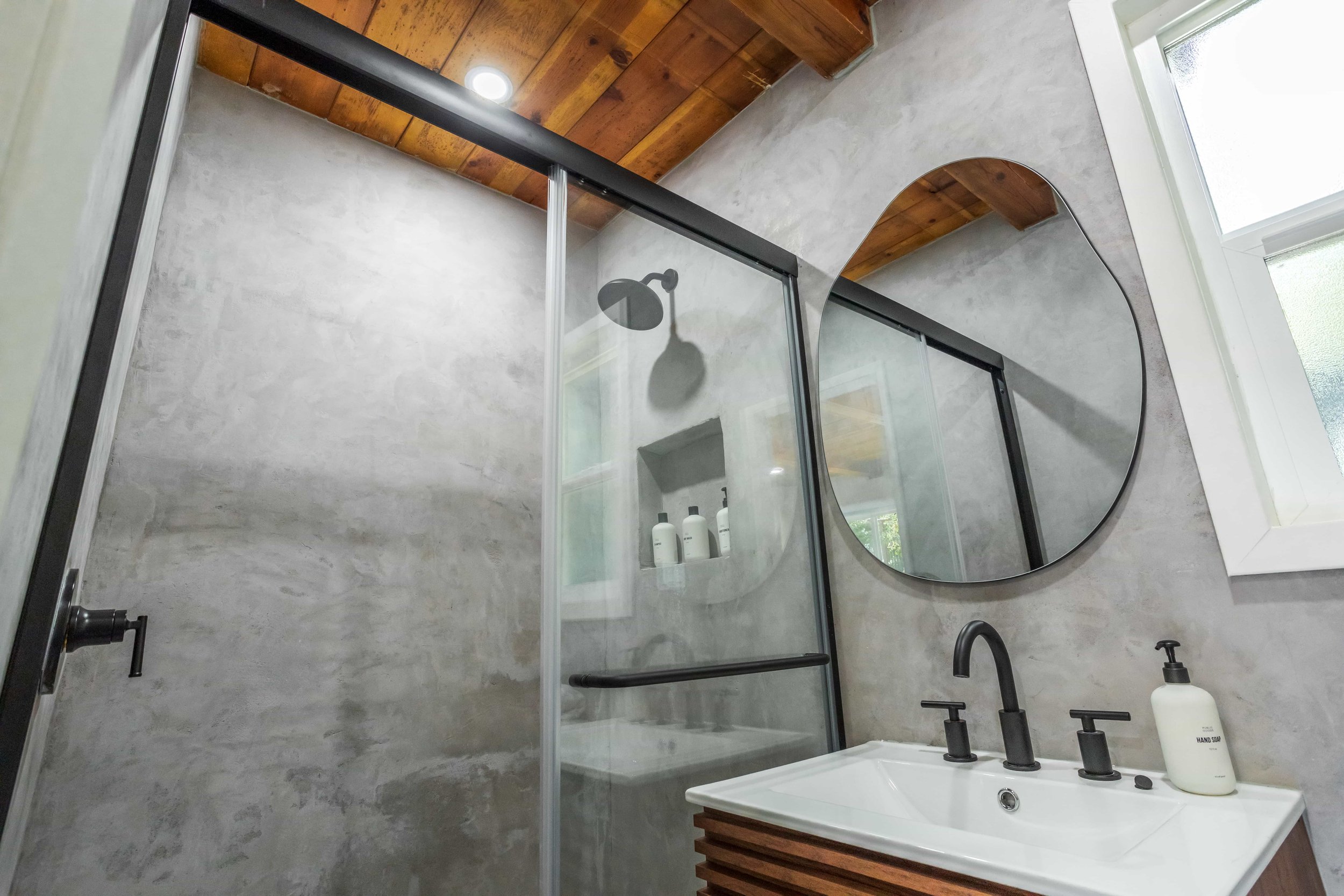 Modern bathroom with concrete walls, a wooden ceiling, a glass shower enclosure with black hardware, a round mirror, and a white sink with black fixtures and a soap dispenser.