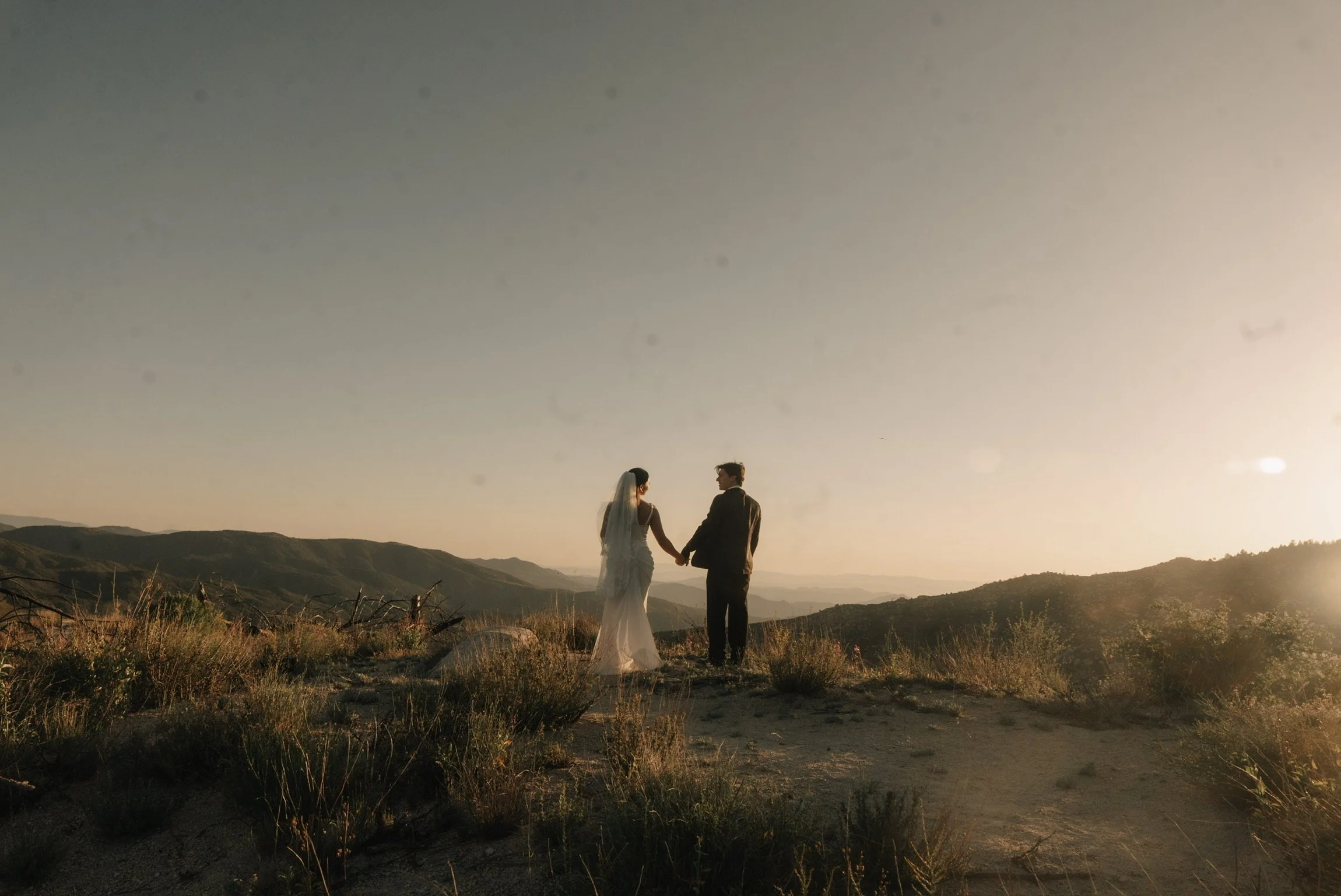 A bride and groom holding hands on a hill at sunset with mountains in the background.