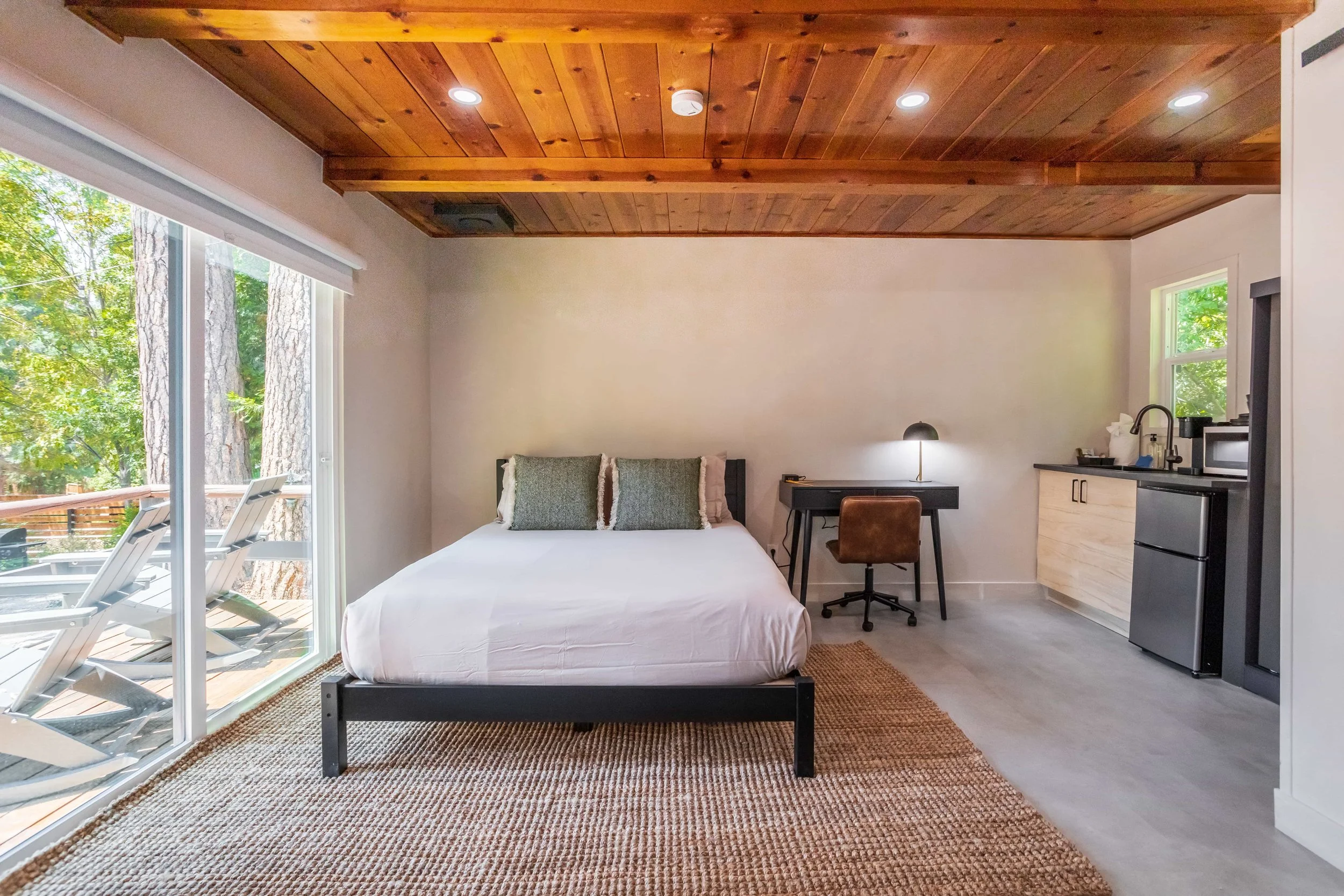 A modern studio apartment with a bed, small kitchen, desk, and sliding glass door leading to a balcony with outdoor chairs.