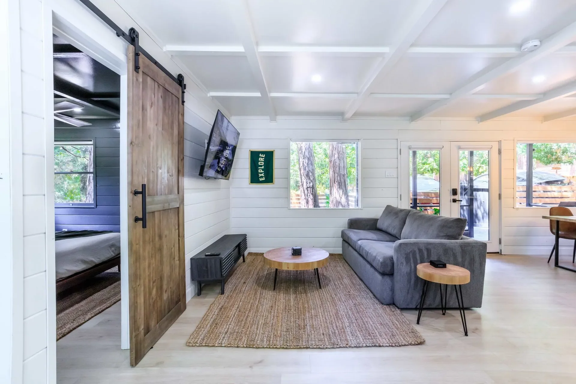 Living room with a gray sofa, wooden coffee table on a rug, TV on the wall, two windows, a door leading outside, and a sliding barn door to a bedroom.