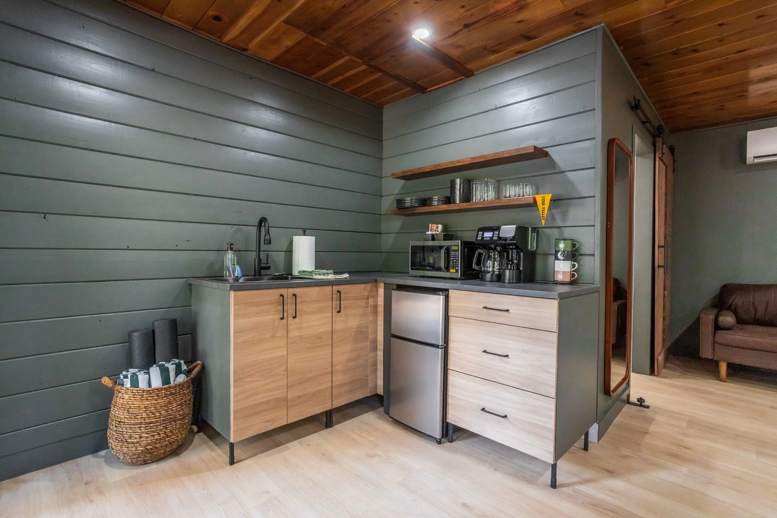 A small kitchen area with wooden cabinets, a mini refrigerator, microwave, coffee maker, and open wooden shelves with dishes. There is a basket with rolled towels or cloths, a paper towel holder, and a sink with a faucet. The walls are painted green,