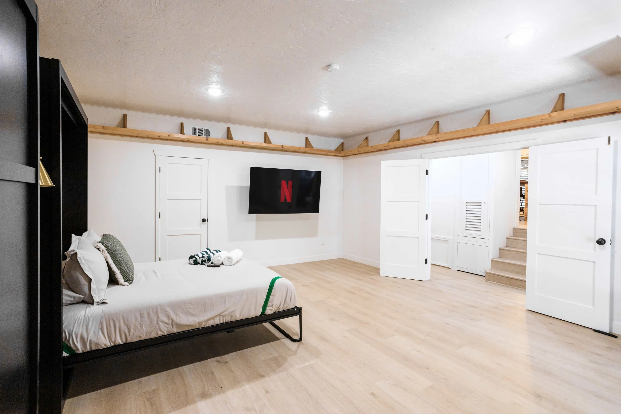 Minimalist bedroom with a bed, throw pillows, towels, and a mounted TV displaying Netflix logo, white walls, light wood flooring, and white doors.