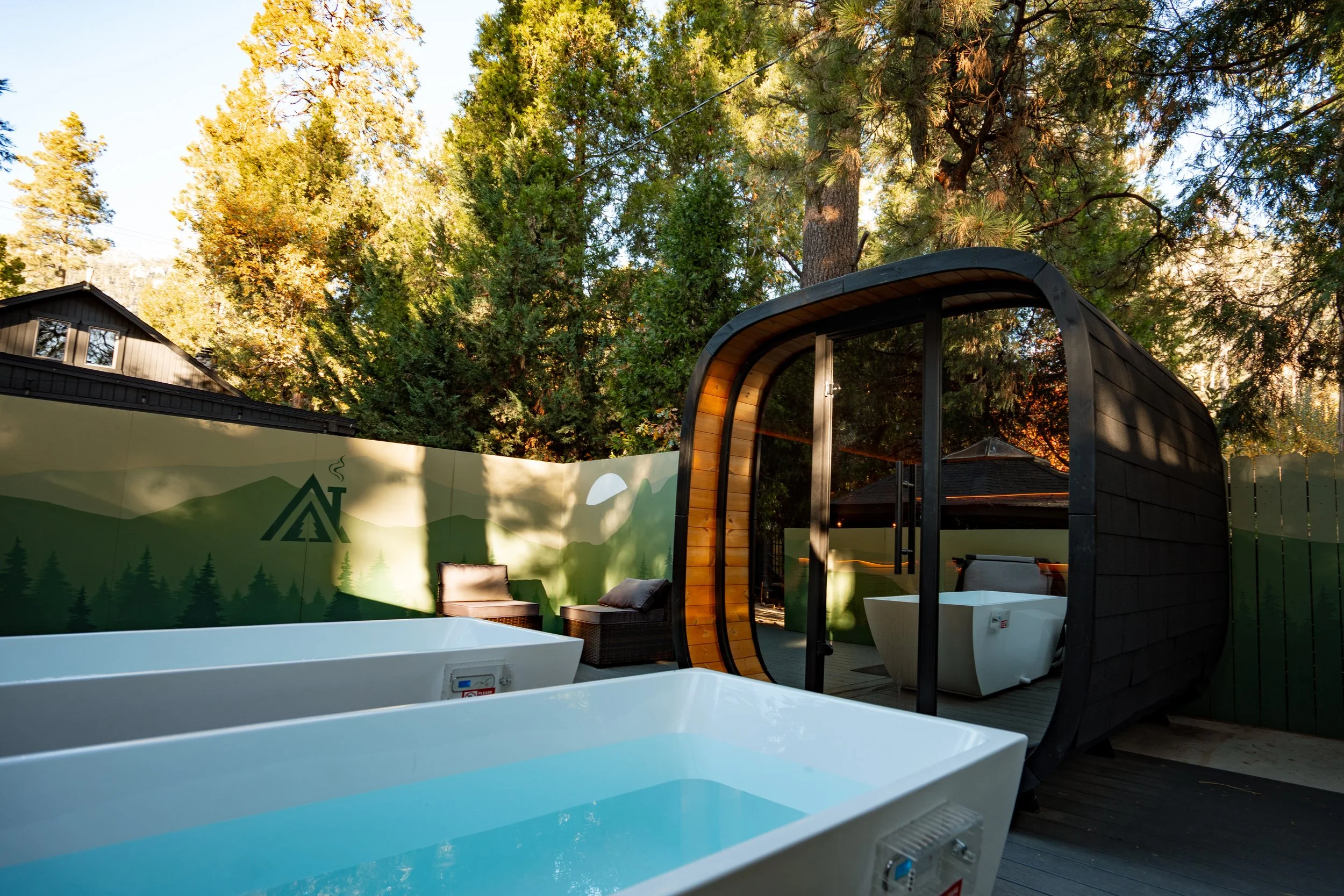 The Pinetree Hotel: Wellness sweat deck : with two cold plunge tubs, a custom sauna, and a seating area surrounded by trees and a painted wooden fence.