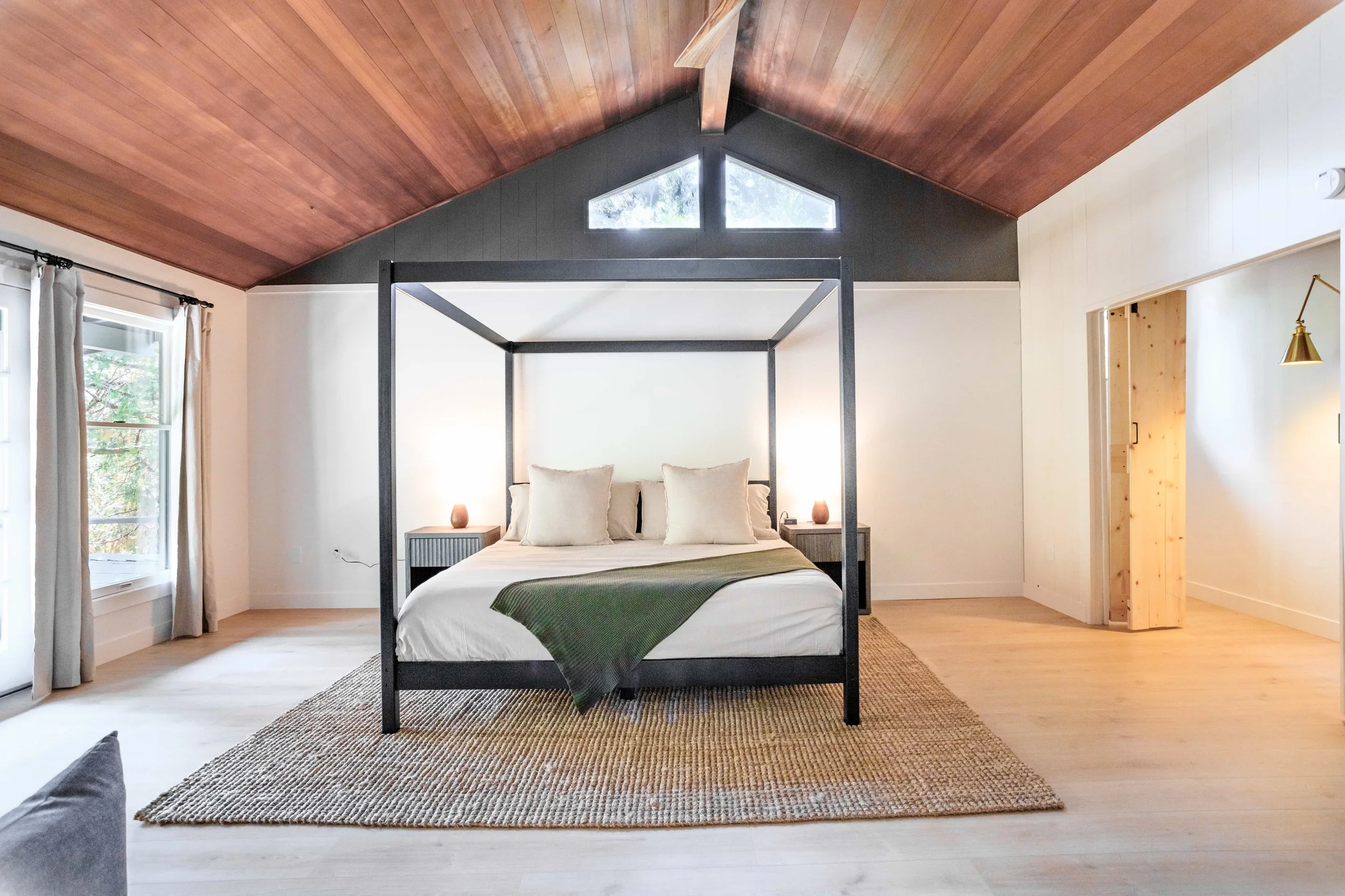 Modern bedroom with a black four-poster bed, white bedding, beige pillows, and a green throw blanket. The room features light wooden floors, a woven rug, and a vaulted wood ceiling. There are windows with light curtains on the left and small bedside 