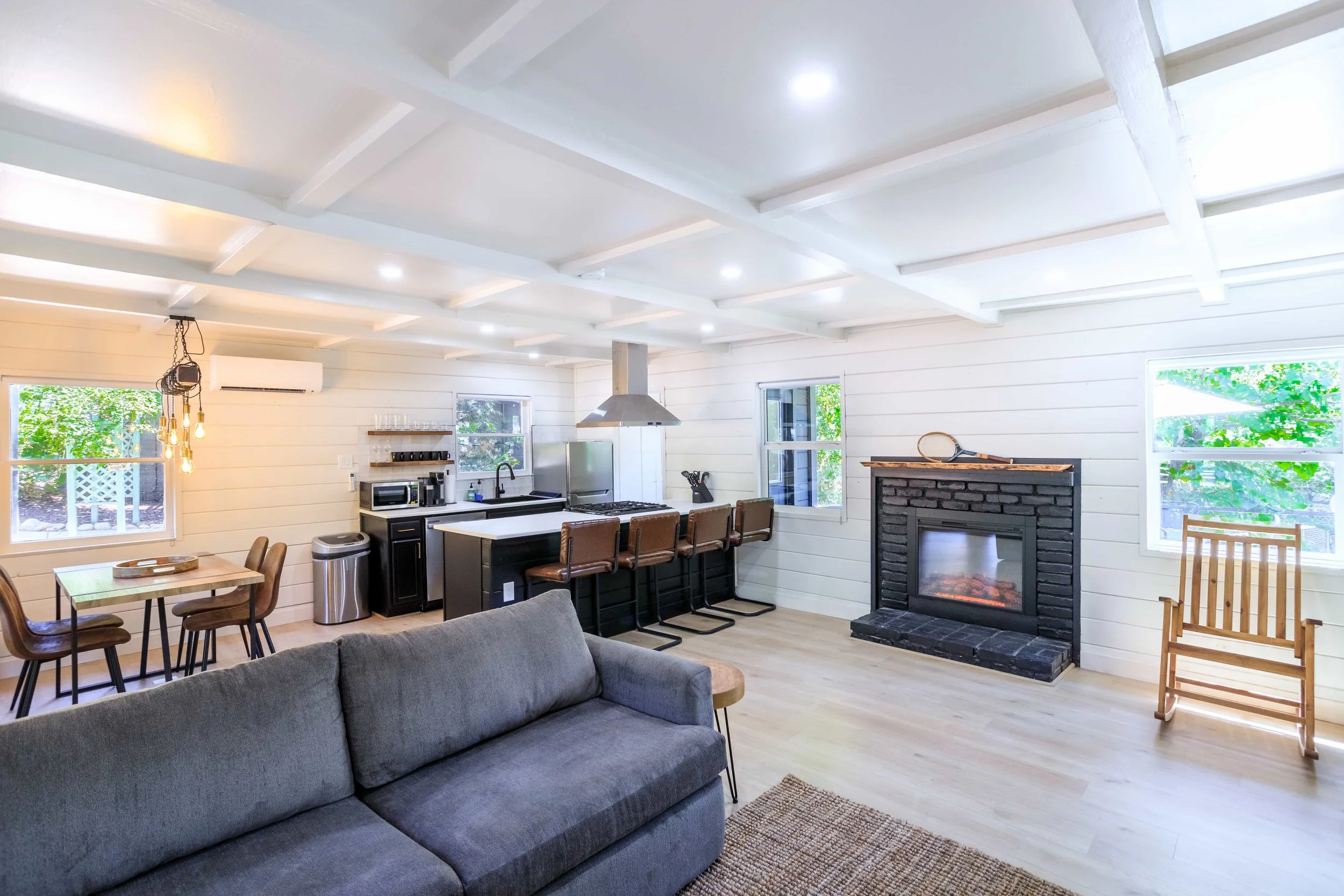 Bright living room with white ceiling and walls, a fireplace, dining area, kitchen with bar stools, and large windows showing green outdoor scenery.