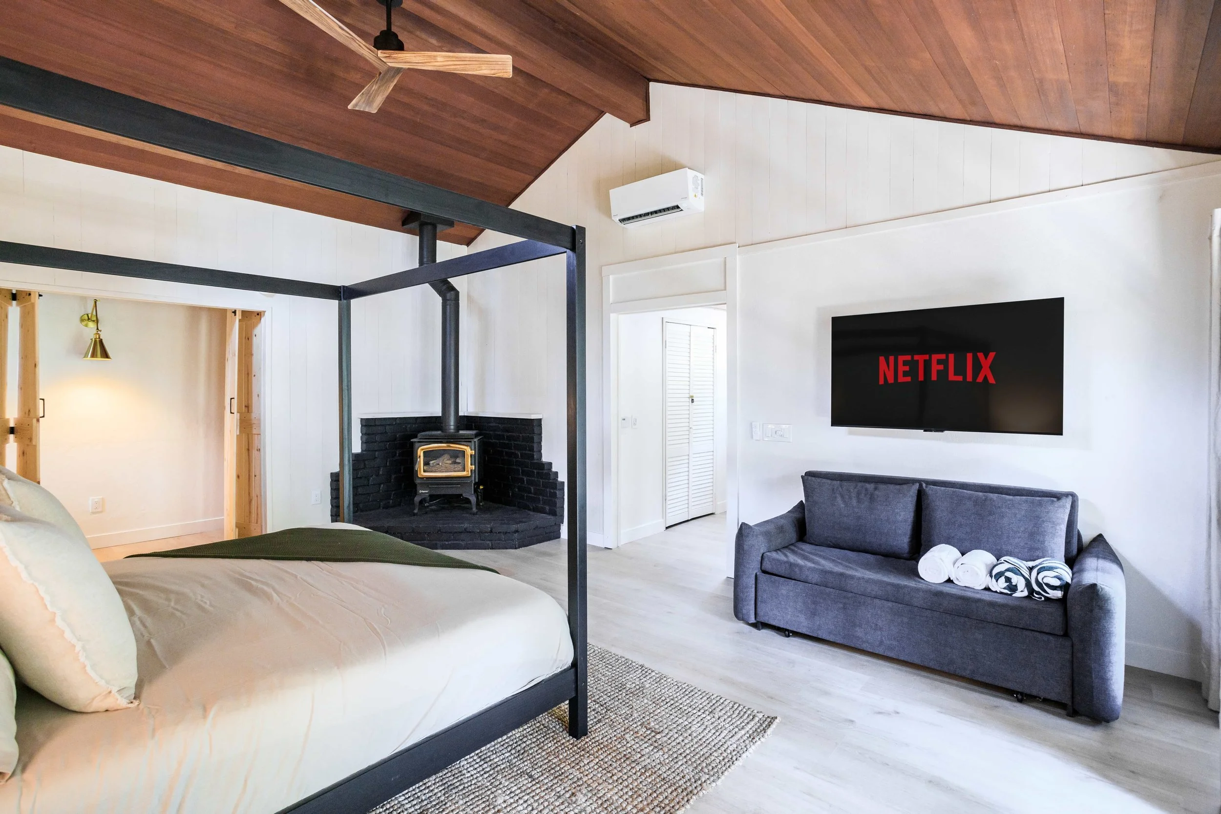 Living room with a bed canopy, a black fireplace, a wall-mounted TV displaying Netflix, and a gray sofa with rolled towels on it.