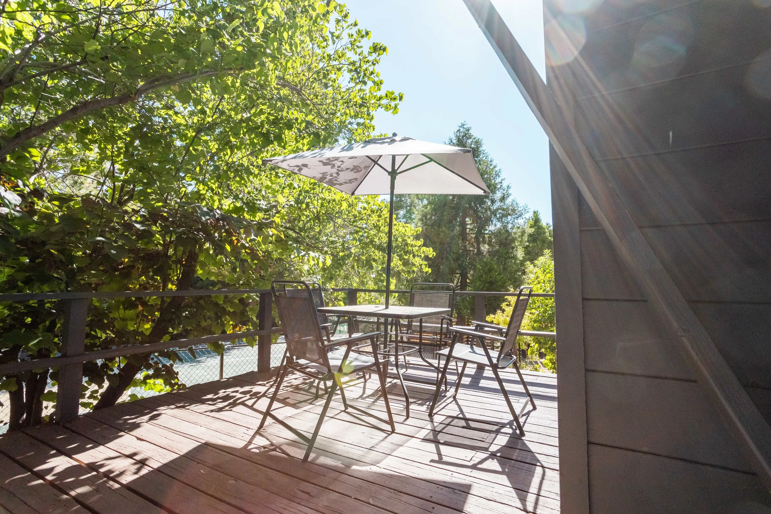 A sunlit outdoor wooden deck with a table, four chairs, and a large white umbrella, surrounded by green trees.