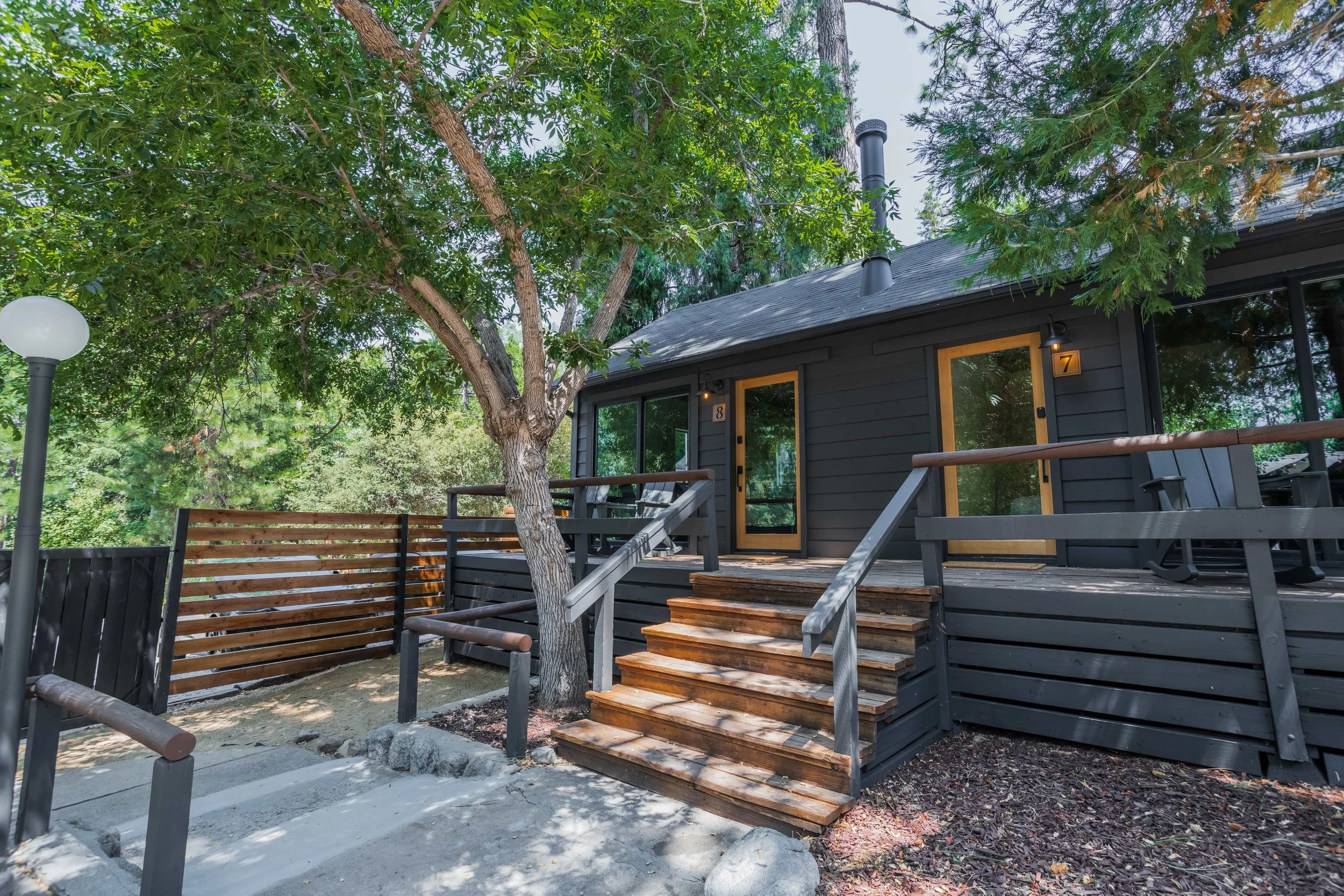 A modern black wooden house with a small front porch, located in a wooded area with large trees. The porch has wooden stairs with black railings, and the house has large glass doors with wood frames. There are small outdoor wall lights and a neighbor