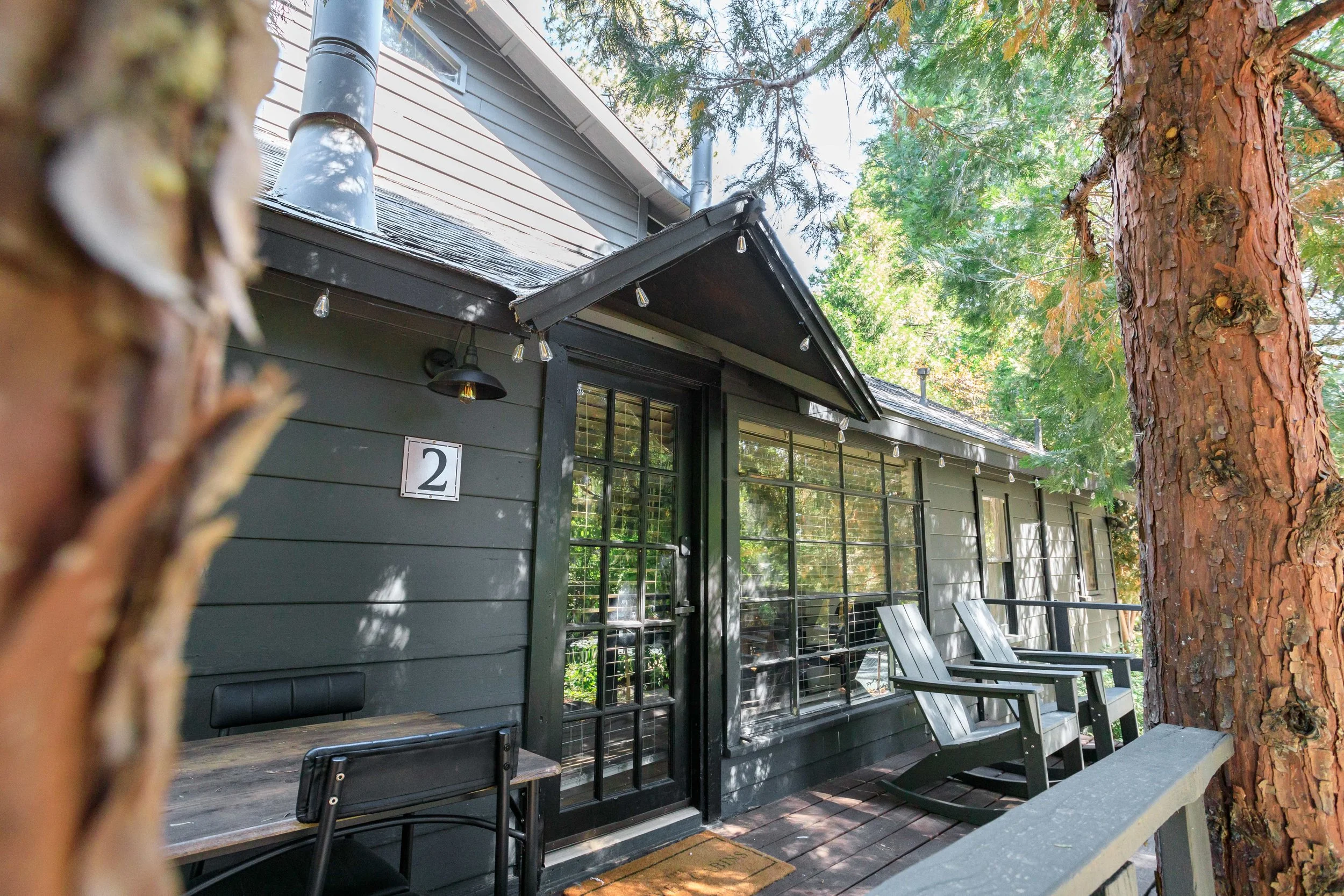 A view of a dark gray house with a porch, featuring a sliding glass door and large windows. There are two black chairs on the porch, and a large tree with a textured trunk is visible on the right. String lights hang along the roof edge, and the backg