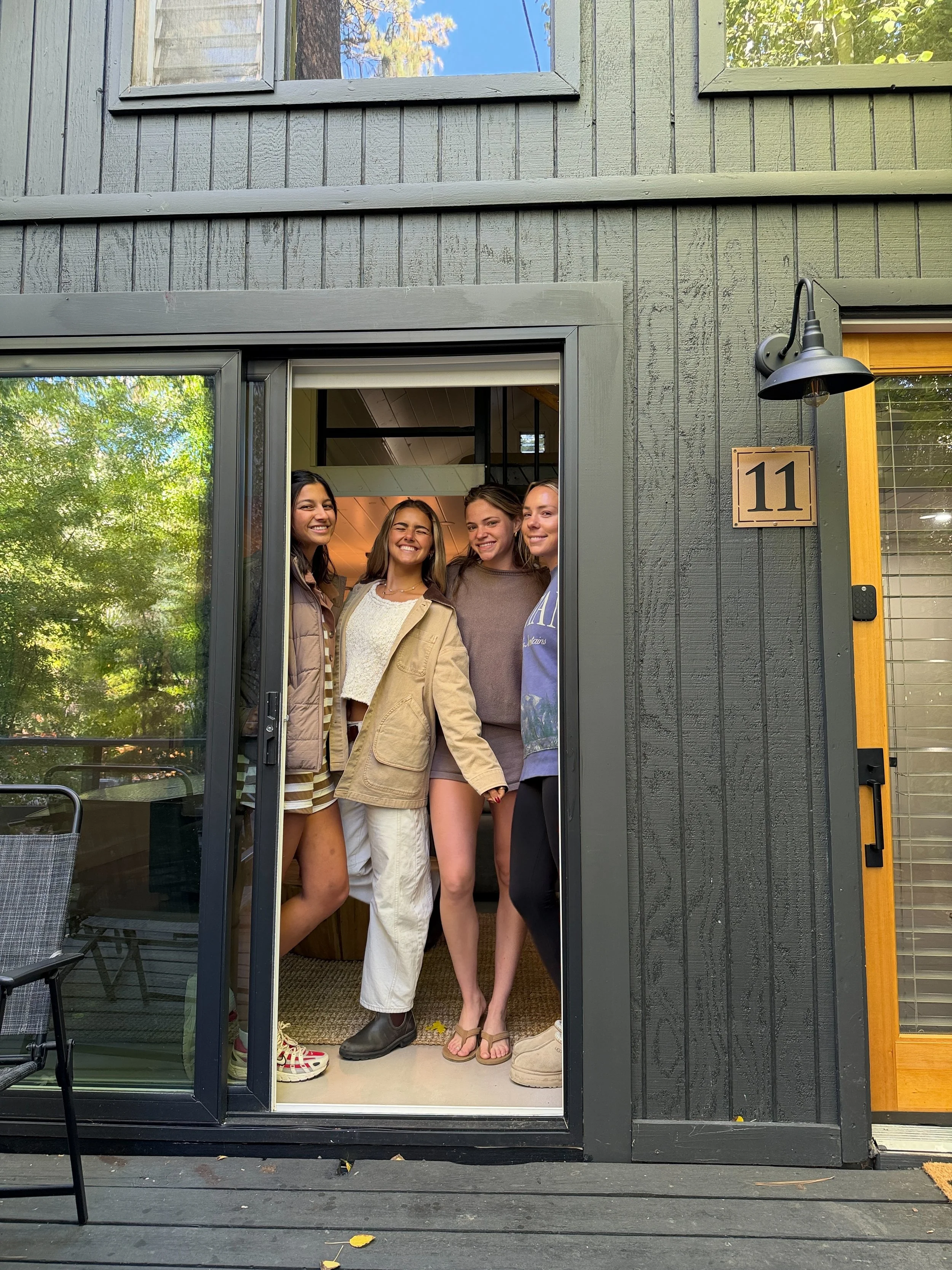 The Pinetree Hotel  cabin 11 guests : Four young women standing together inside a house, visible through the open glass door, smiling at the camera. The house has black exterior walls, a house number '11', and a light fixture above the door.