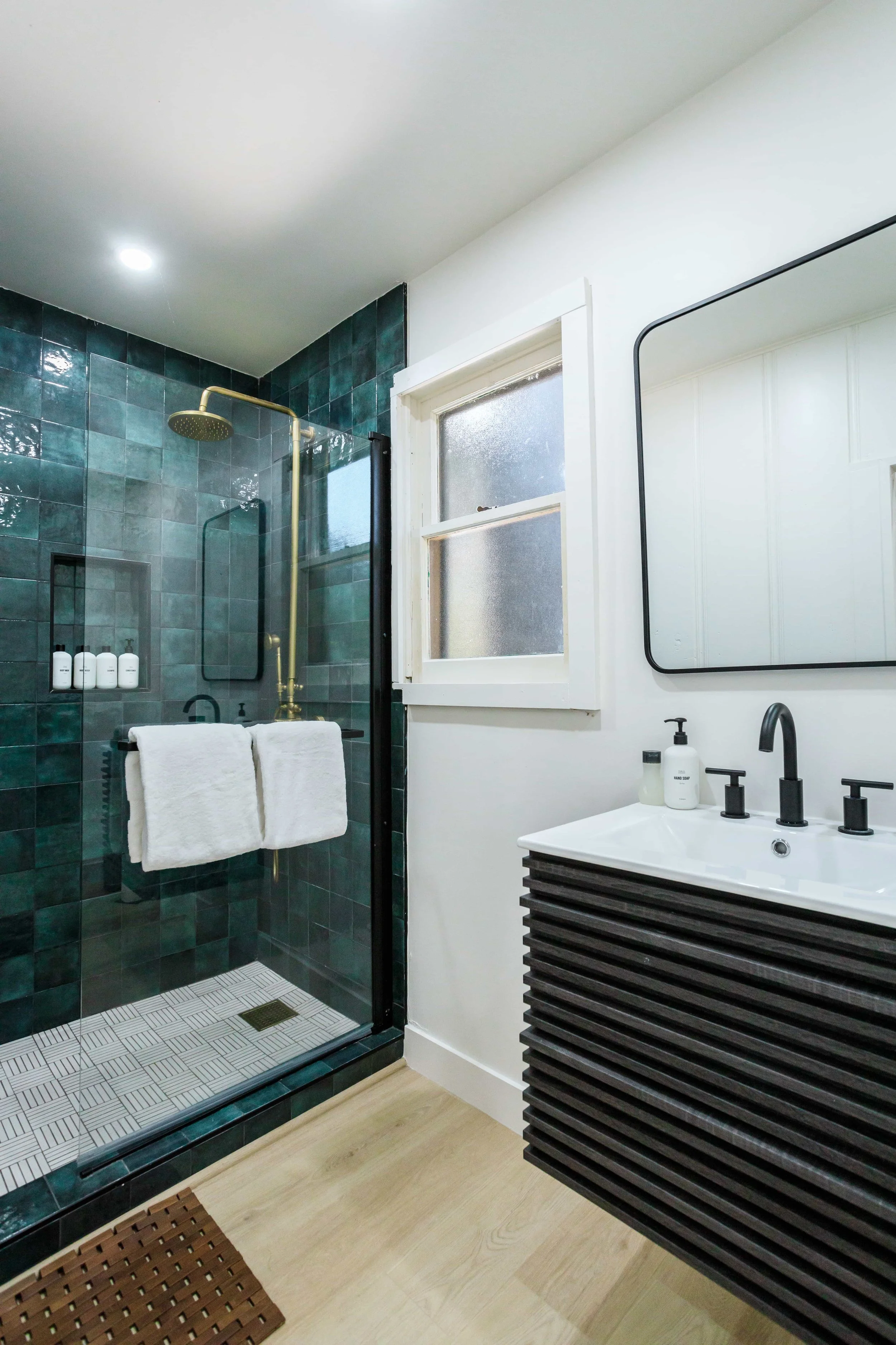 Modern bathroom with a walk-in shower featuring dark green tiles, a brass showerhead, and a glass door. A black-framed mirror hangs above a black slatted vanity with a white sink, black faucet, and soap dispensers. White towels are hanging on the sho