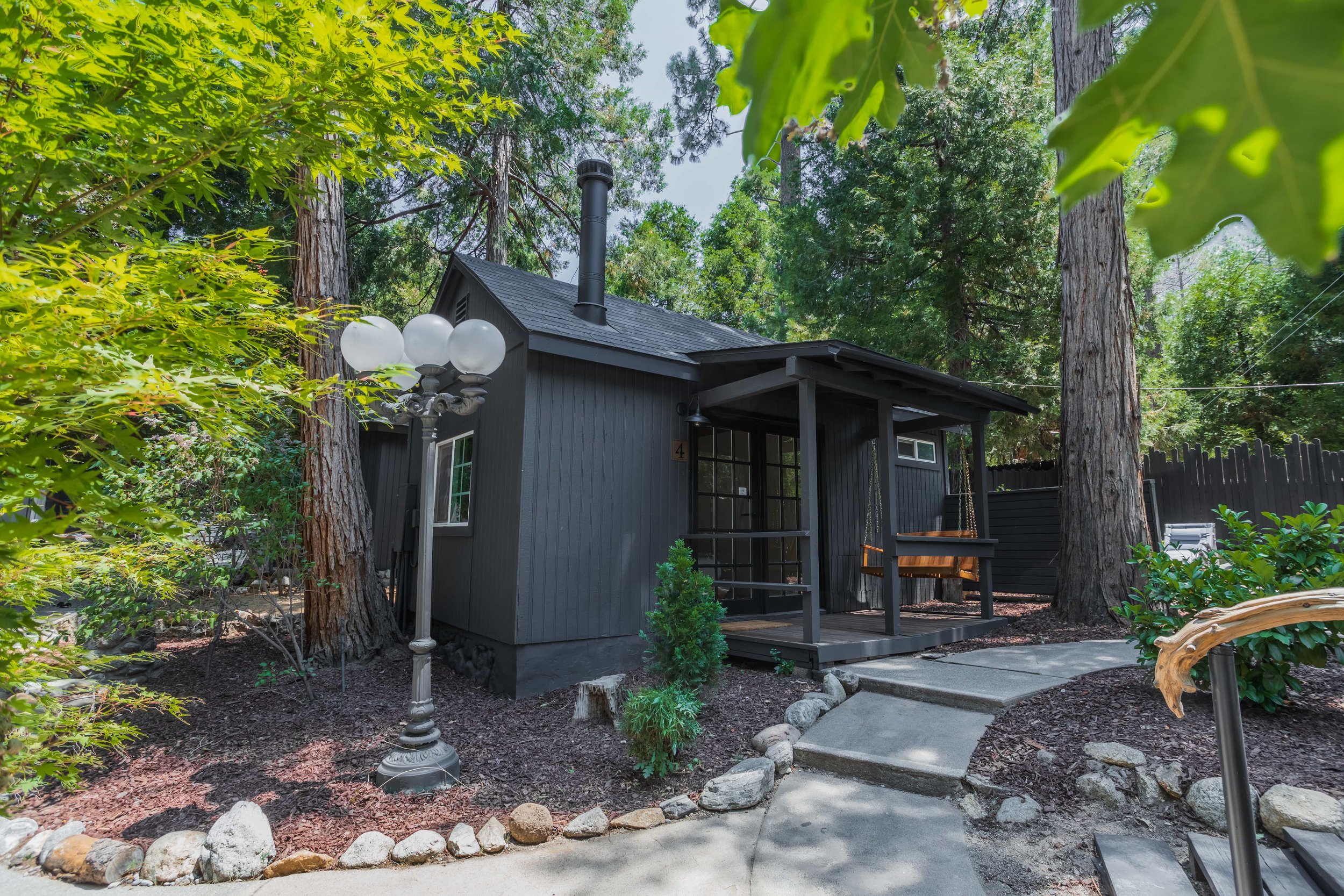 A black wooden house with a porch and swing surrounded by tall trees and lush greenery.