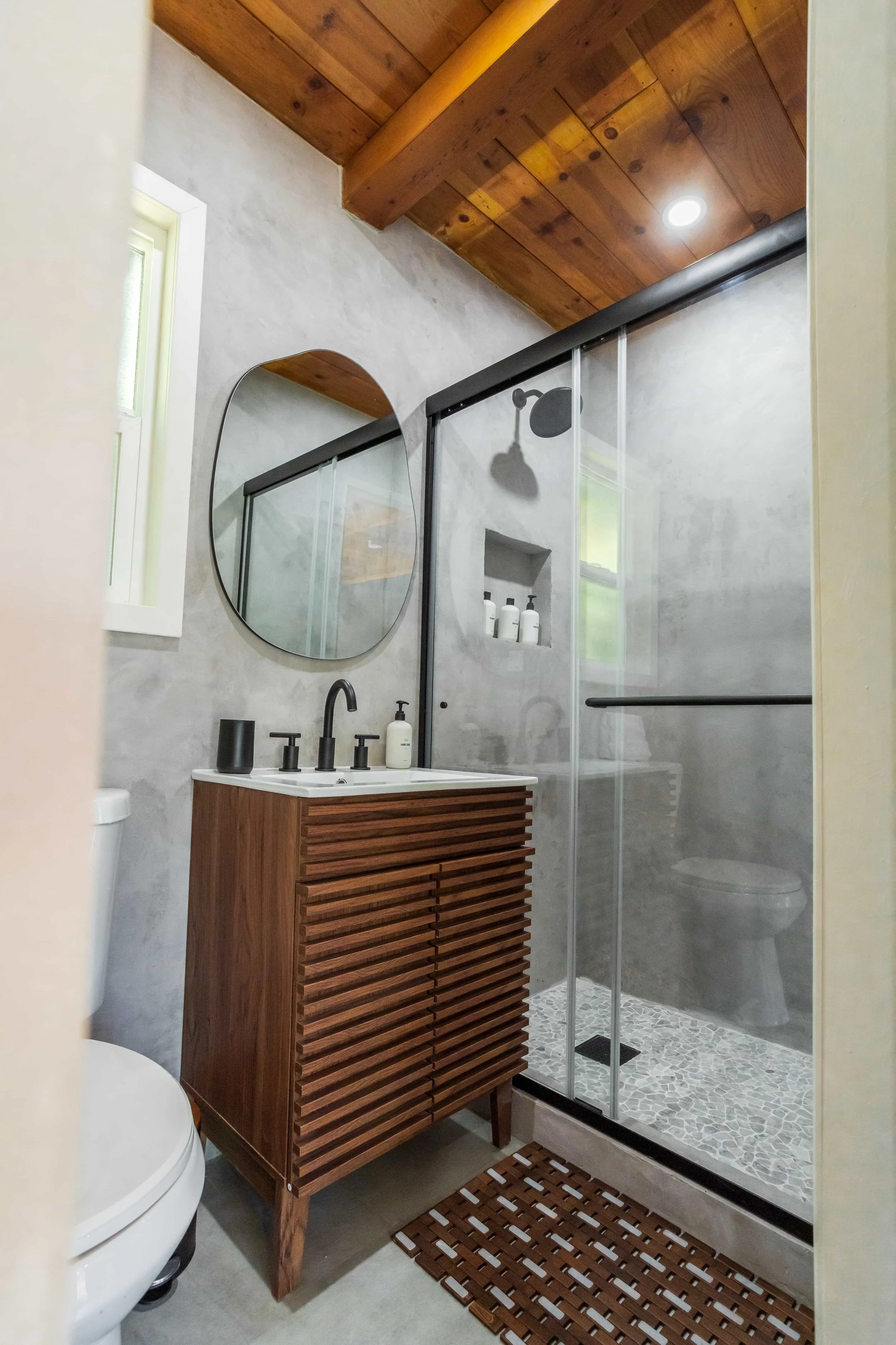 A modern bathroom featuring a wooden vanity with a white countertop, a round mirror, a glass shower enclosure with pebble tile flooring, a rainfall showerhead, and a wooden ceiling with recessed lighting.