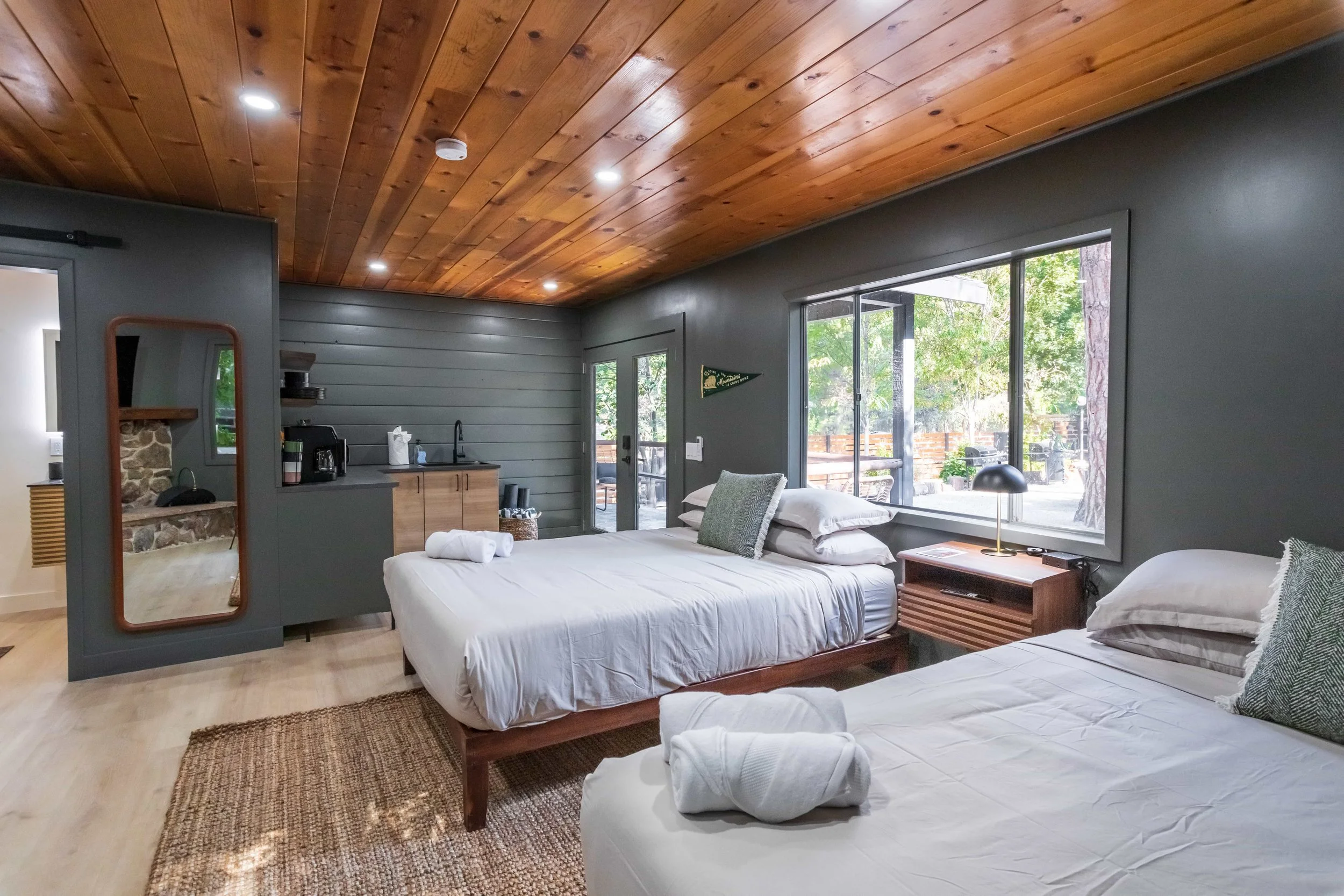 A cozy bedroom with two beds, a wooden ceiling, and large windows showing green trees outside. There is a nightstand with a lamp and a small kitchenette with a sink and coffee maker in the corner.