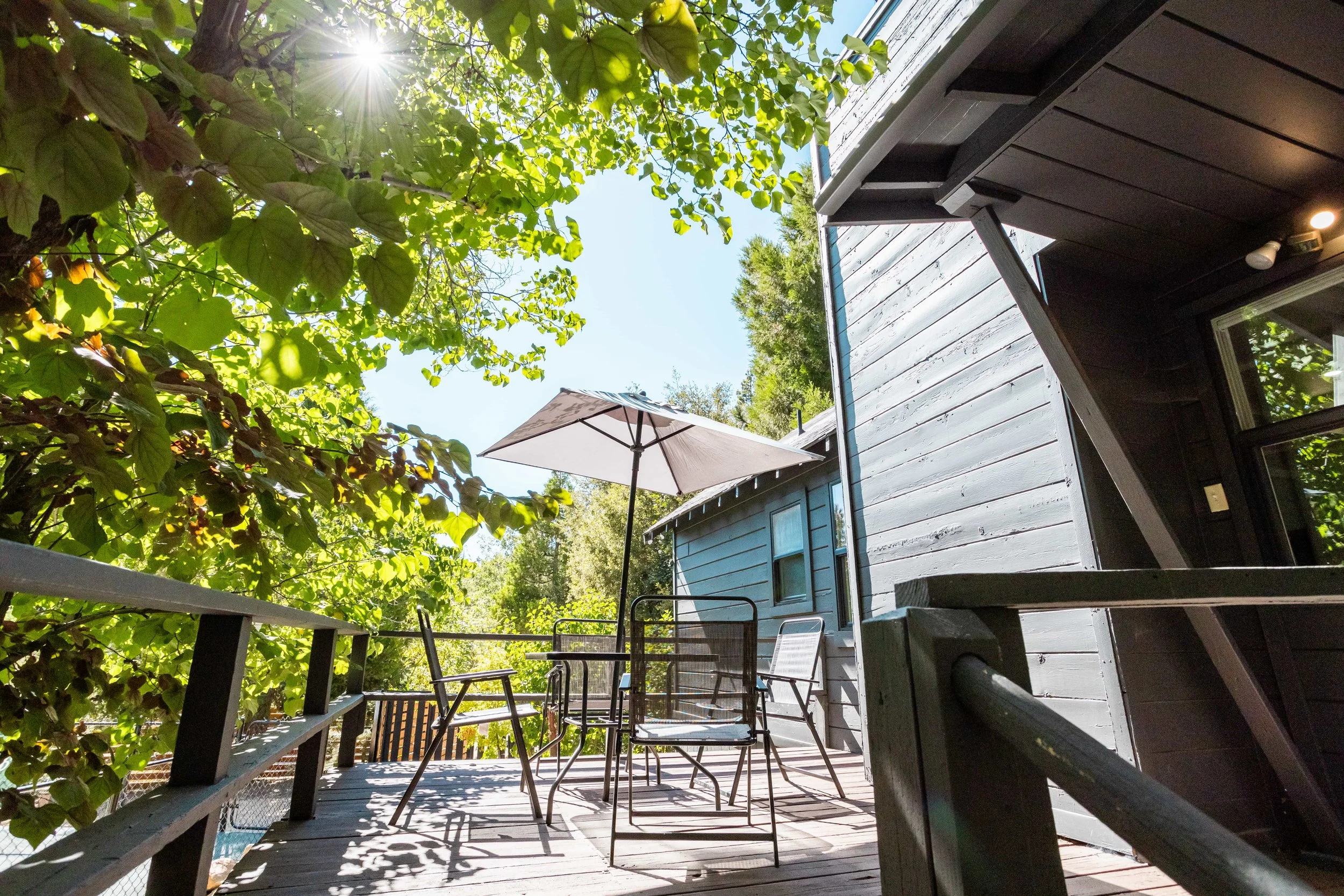View of an outdoor wooden deck with patio furniture including a table, chairs, and an umbrella, surrounded by green foliage with sunlight shining through the leaves.