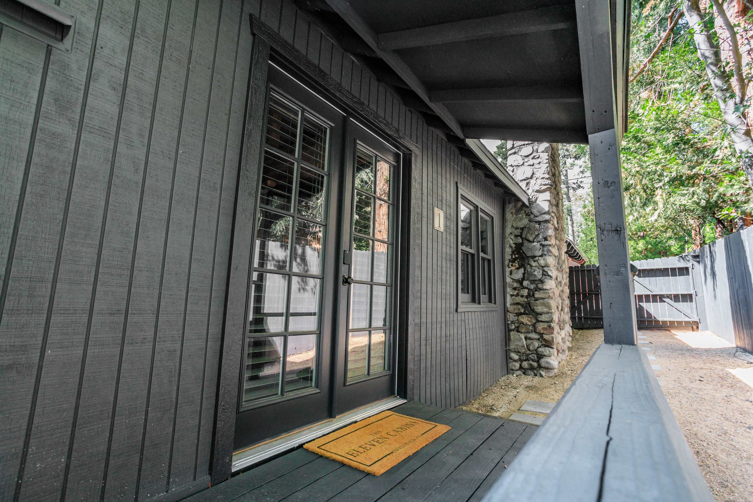 Front porch of a house with black wooden siding, glass-paneled doors, a yellow welcome mat, and a gravel walkway.