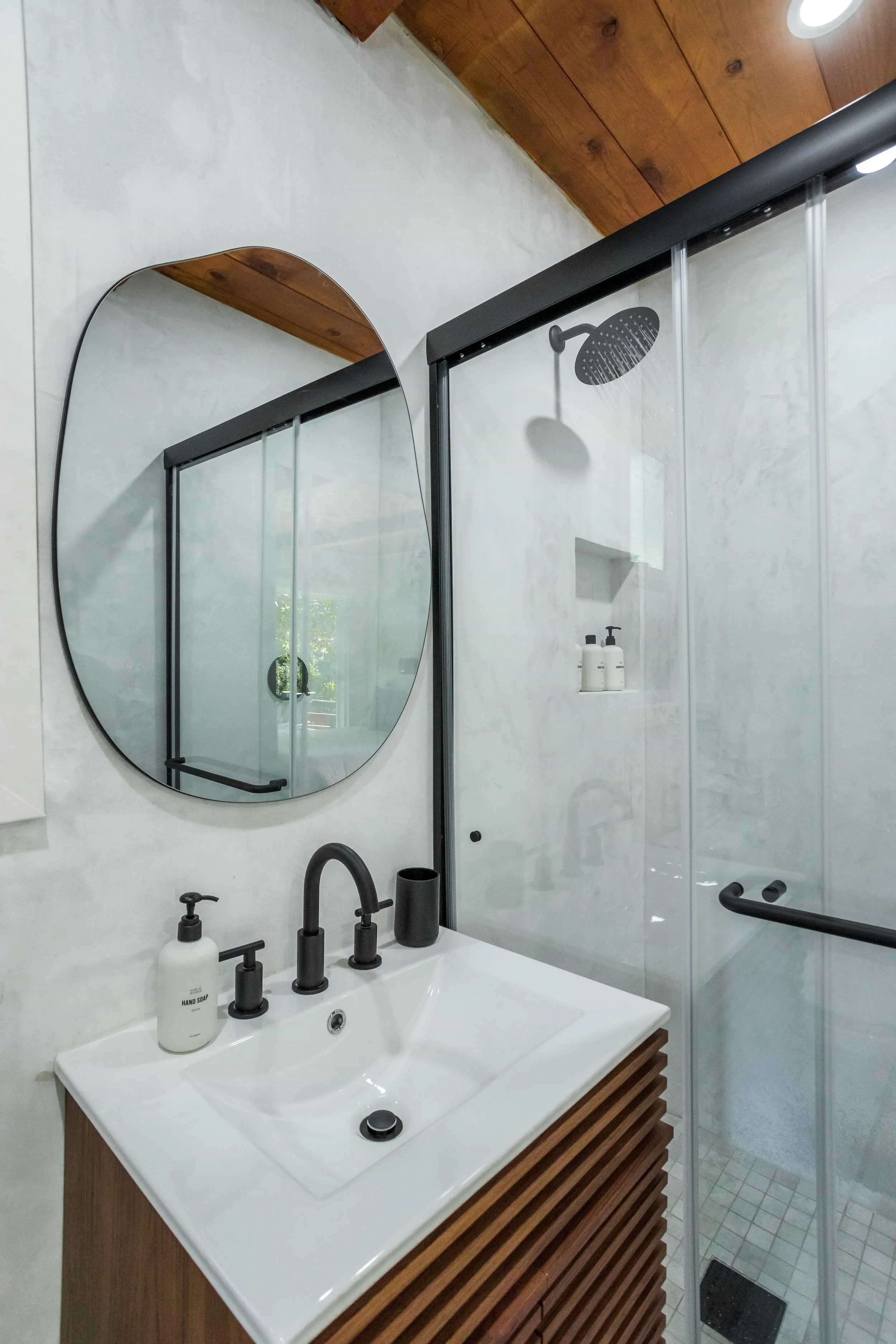 Modern bathroom with a white sink, black fixtures, a round mirror, a glass shower with black hardware, and a wooden ceiling.