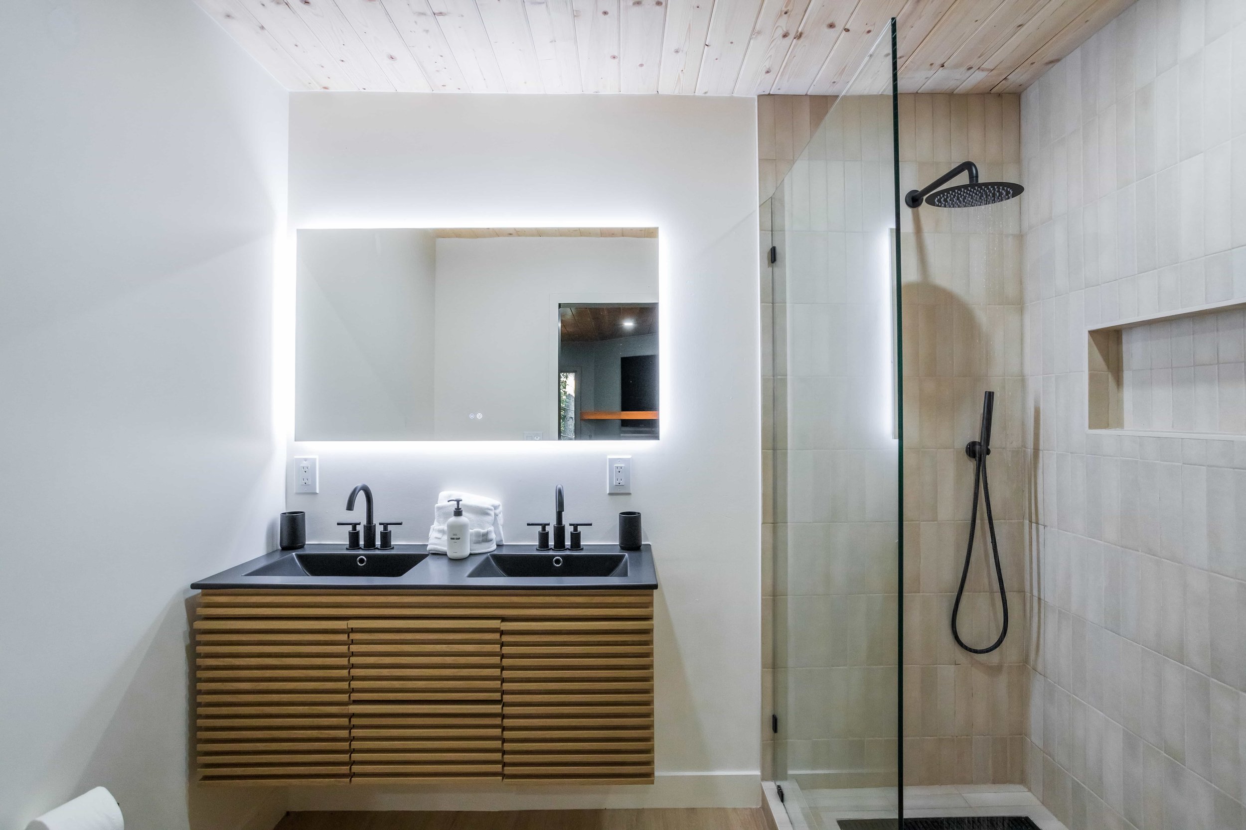 Modern bathroom with a double sink vanity, illuminated mirror, and a walk-in shower with a glass door, black fixtures, and beige tiled walls.