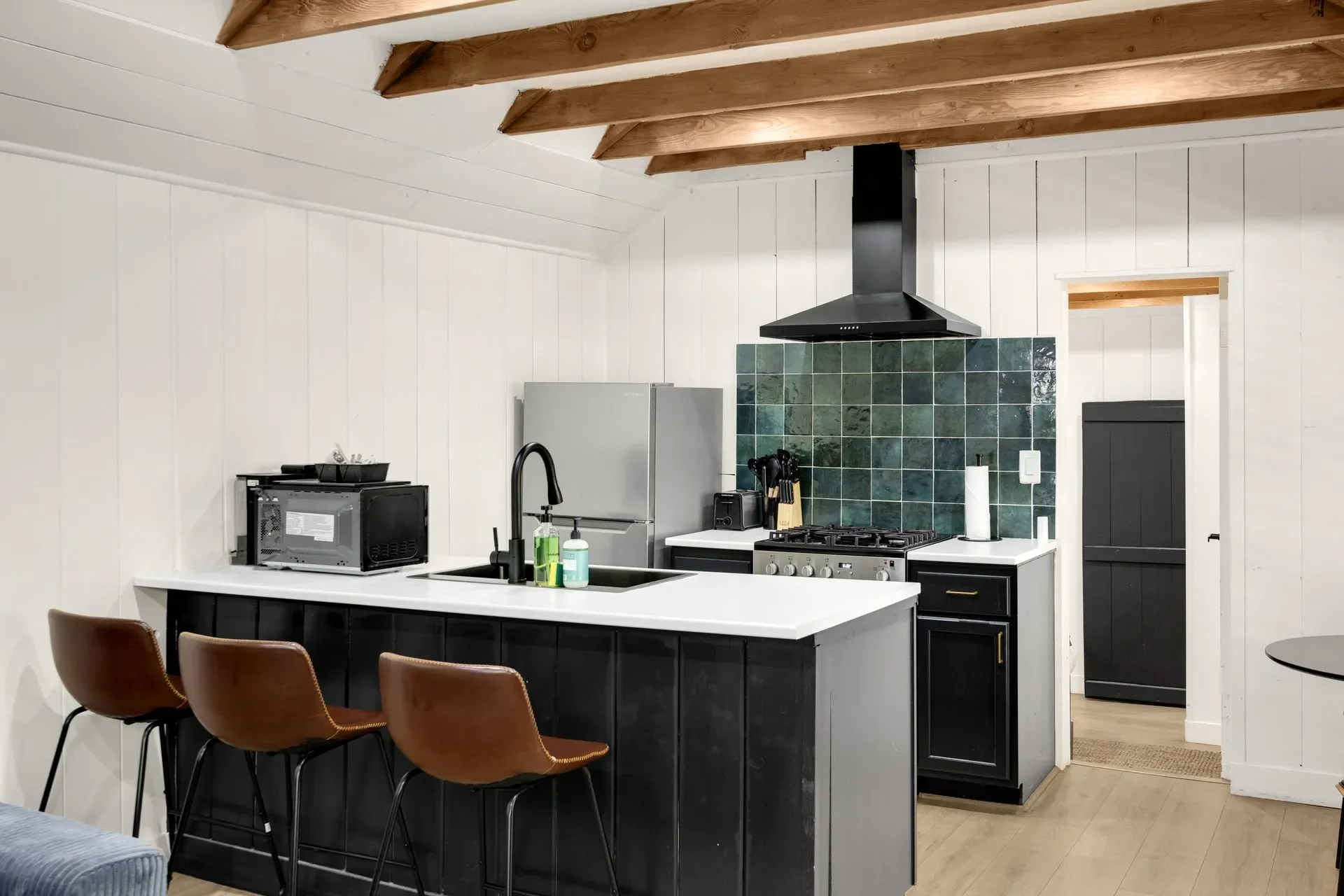 Modern kitchen with black lower cabinets, white countertop, green tiled backsplash, stainless steel refrigerator, and black range hood. Three brown chairs are at the kitchen island. White walls with wood ceiling beams.