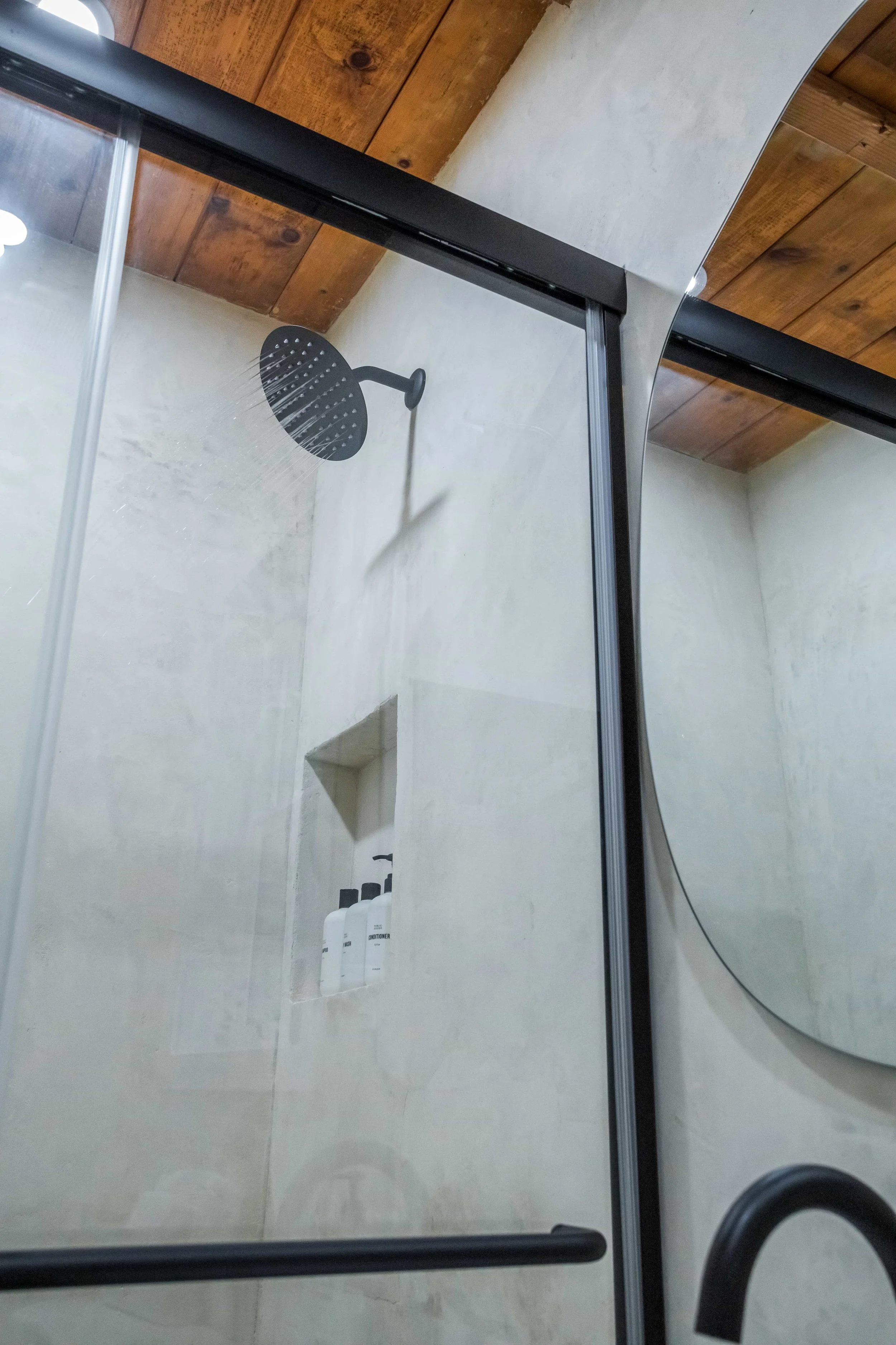 Close-up of a shower with a black rain showerhead mounted on a white wall, behind a glass door with black framing, and a built-in niche holding multiple bottles of toiletries, with wooden ceiling above.