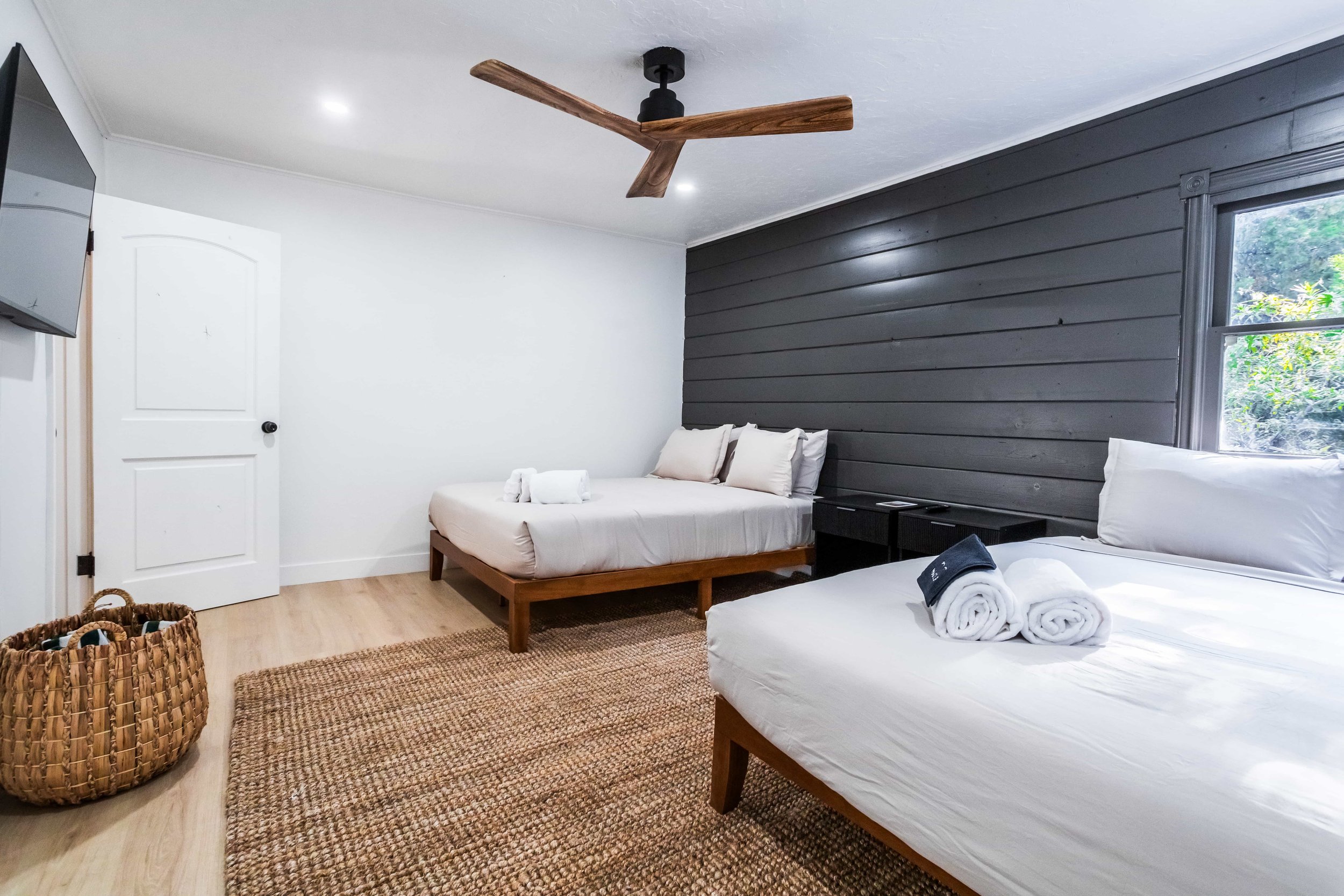 The Pinetree hotel cabin 1 - guest room . A bedroom with two beds, one with white linens and the other with beige linens, a dark gray wooden accent wall, a window, a ceiling fan, a TV on the wall, a woven basket, and a brown area rug.