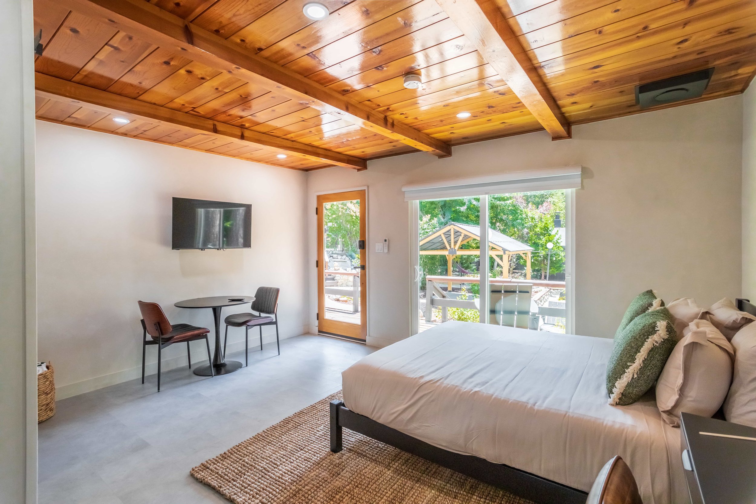 A bedroom with a wooden ceiling, a bed with white linens, a small table with two chairs, a wall-mounted TV, and a sliding glass door leading to a balcony with outdoor furniture and green trees.