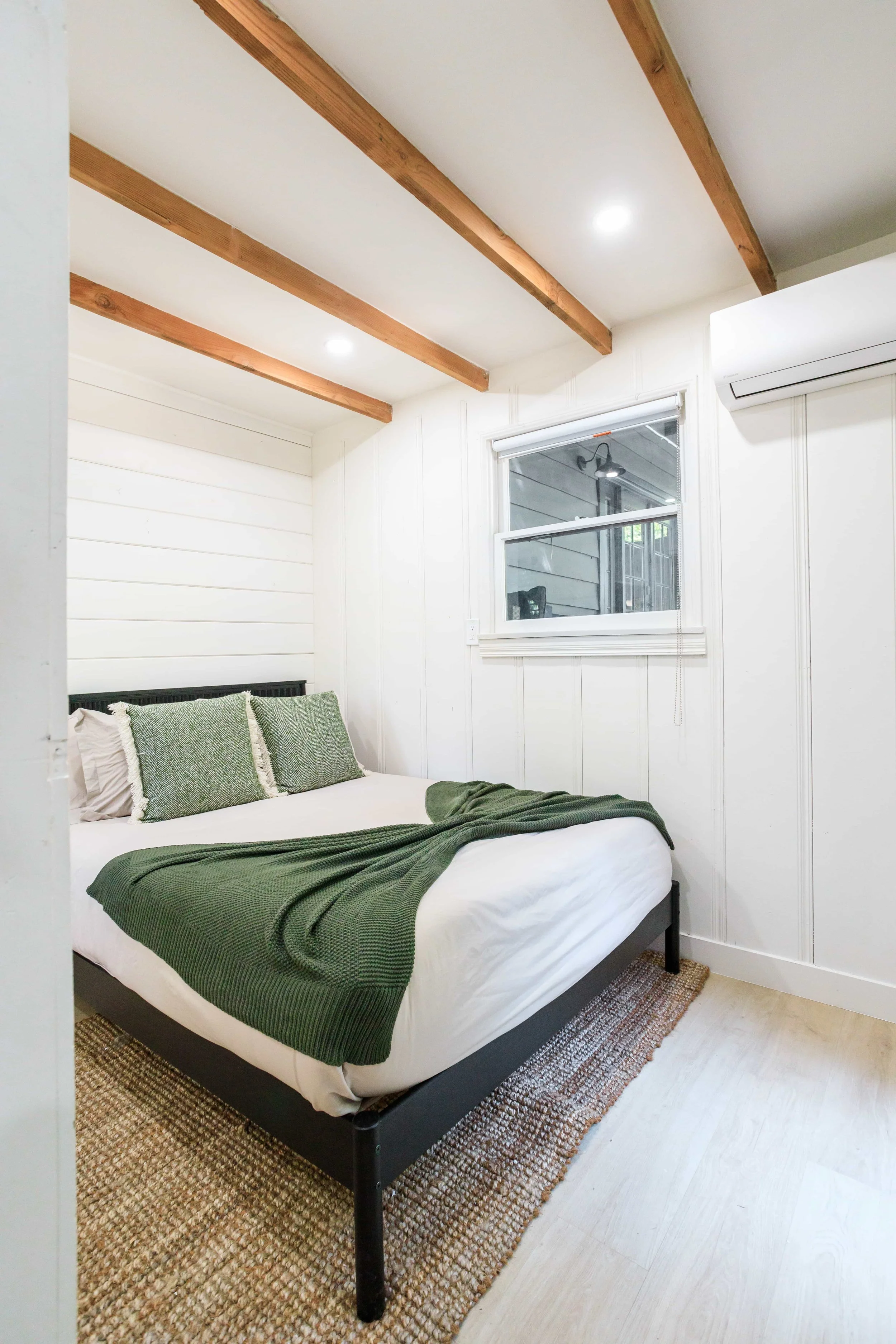 A minimalist bedroom with a black bed frame, white bedding, a green blanket, and matching green pillows. The room has white paneled walls, a window, and wooden ceiling beams.