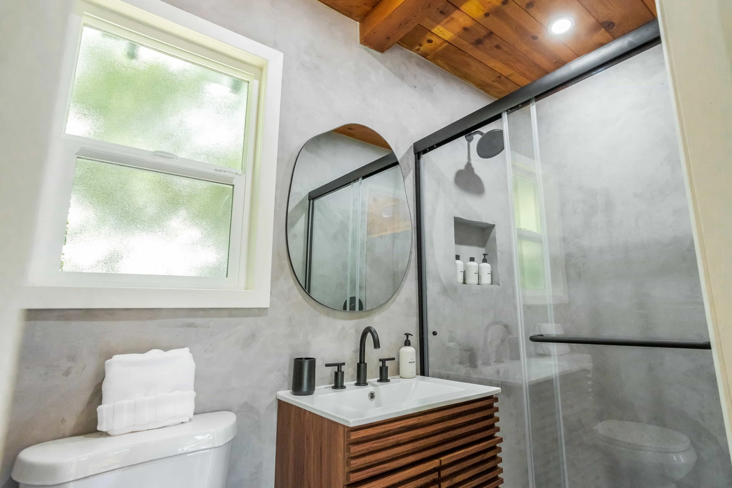 A modern bathroom with a frosted window, a round mirror, a wooden vanity with a white sink, a black faucet, a toothbrush holder, lotion bottle, a shower with frosted glass door, built-in shelves with shampoo bottles, and a wood-paneled ceiling.