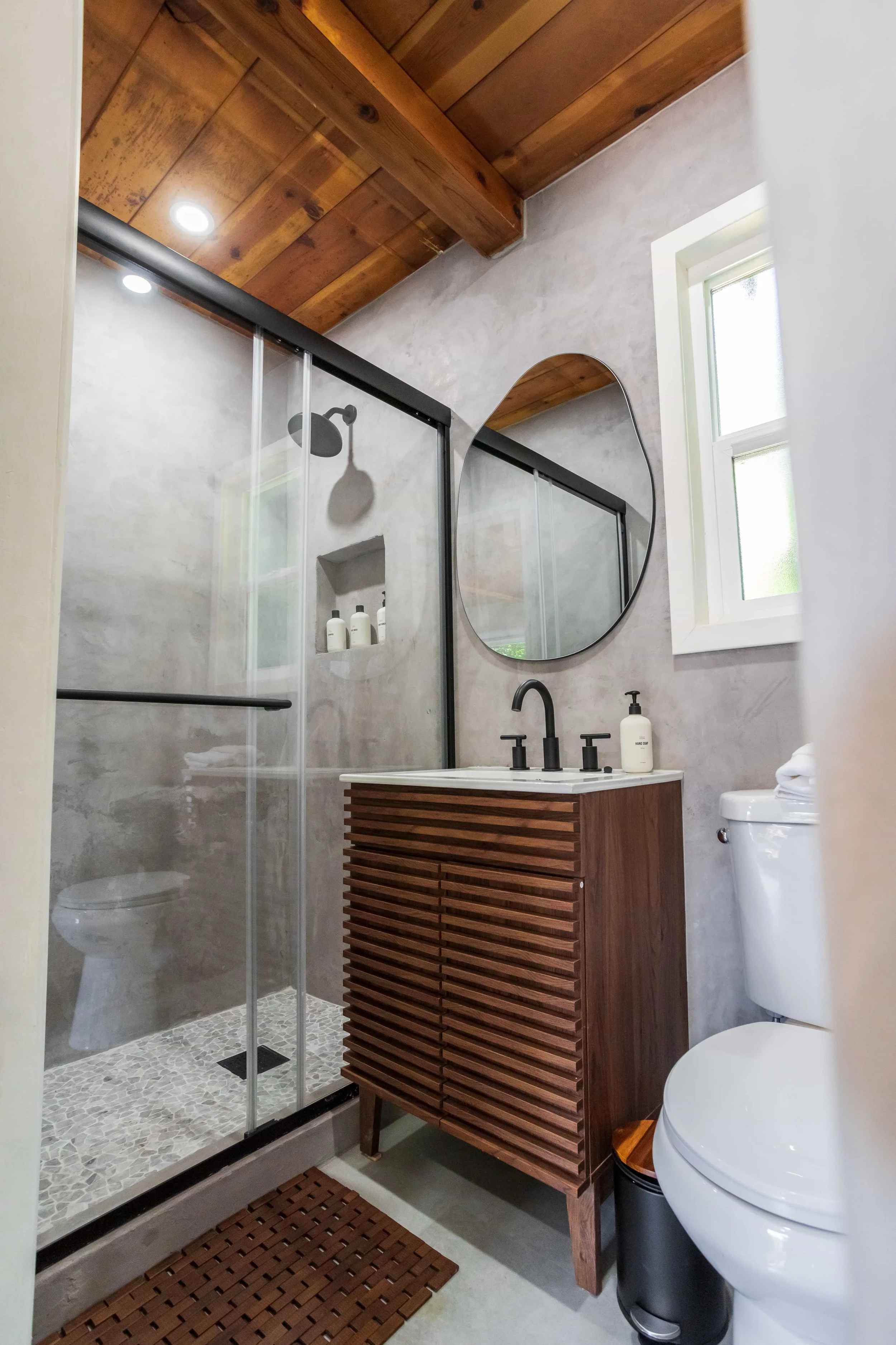 Modern bathroom with walk-in shower, wooden vanity with a circular mirror, a window, and a toilet.