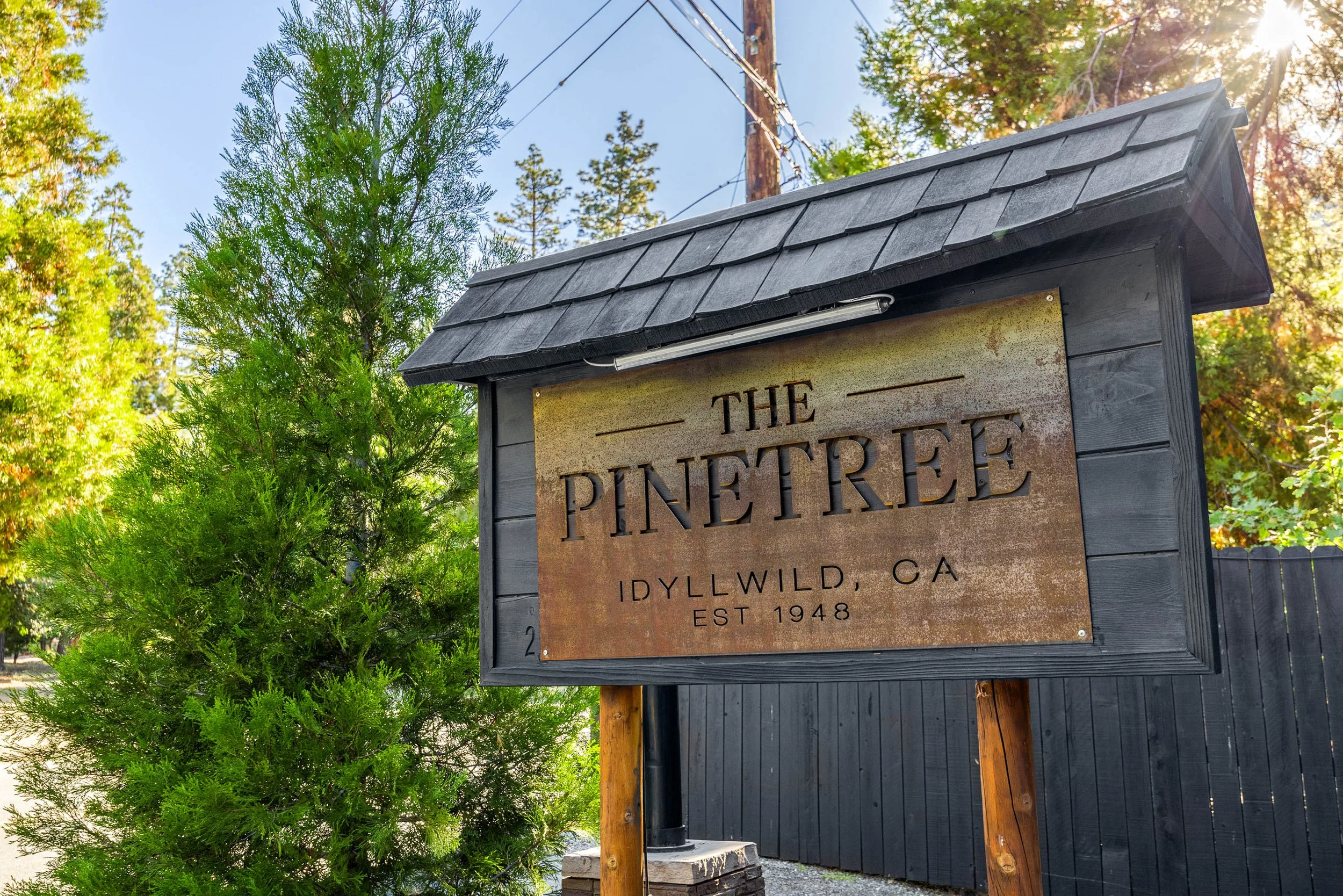 A sign for The Pinetree located in Idyllwild, California, with a rustic wooden design, surrounded by trees and sunlight.