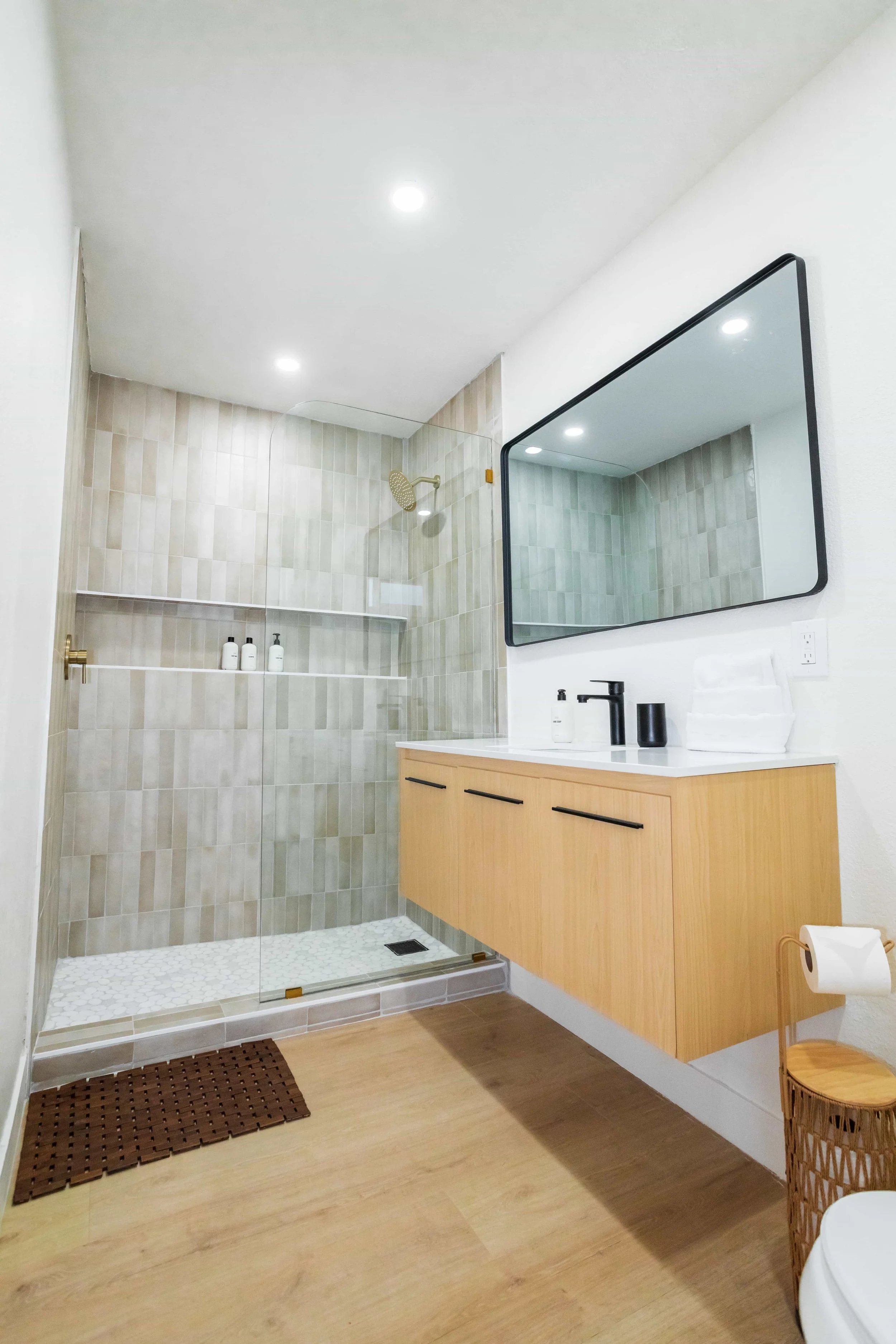 Modern bathroom with a walk-in shower, wooden vanity with black handles, rectangular mirror, white countertop, black faucet, and minimal decor.