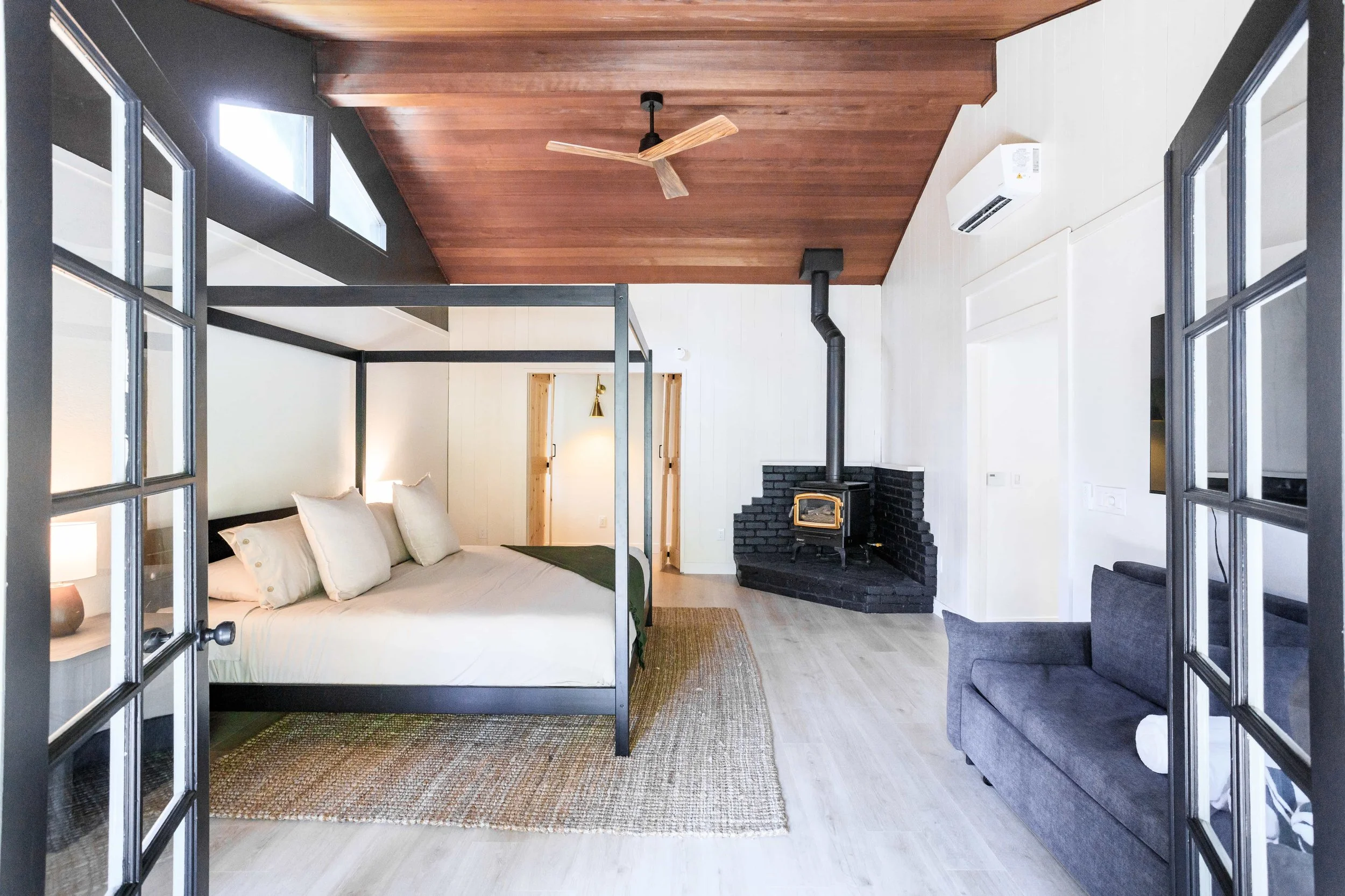 Interior of a modern bedroom with a black metal four poster bed with white bedding and pillows, a gray sofa, a brick fireplace with a wood stove, wood ceiling, light-colored flooring, and double glass doors.