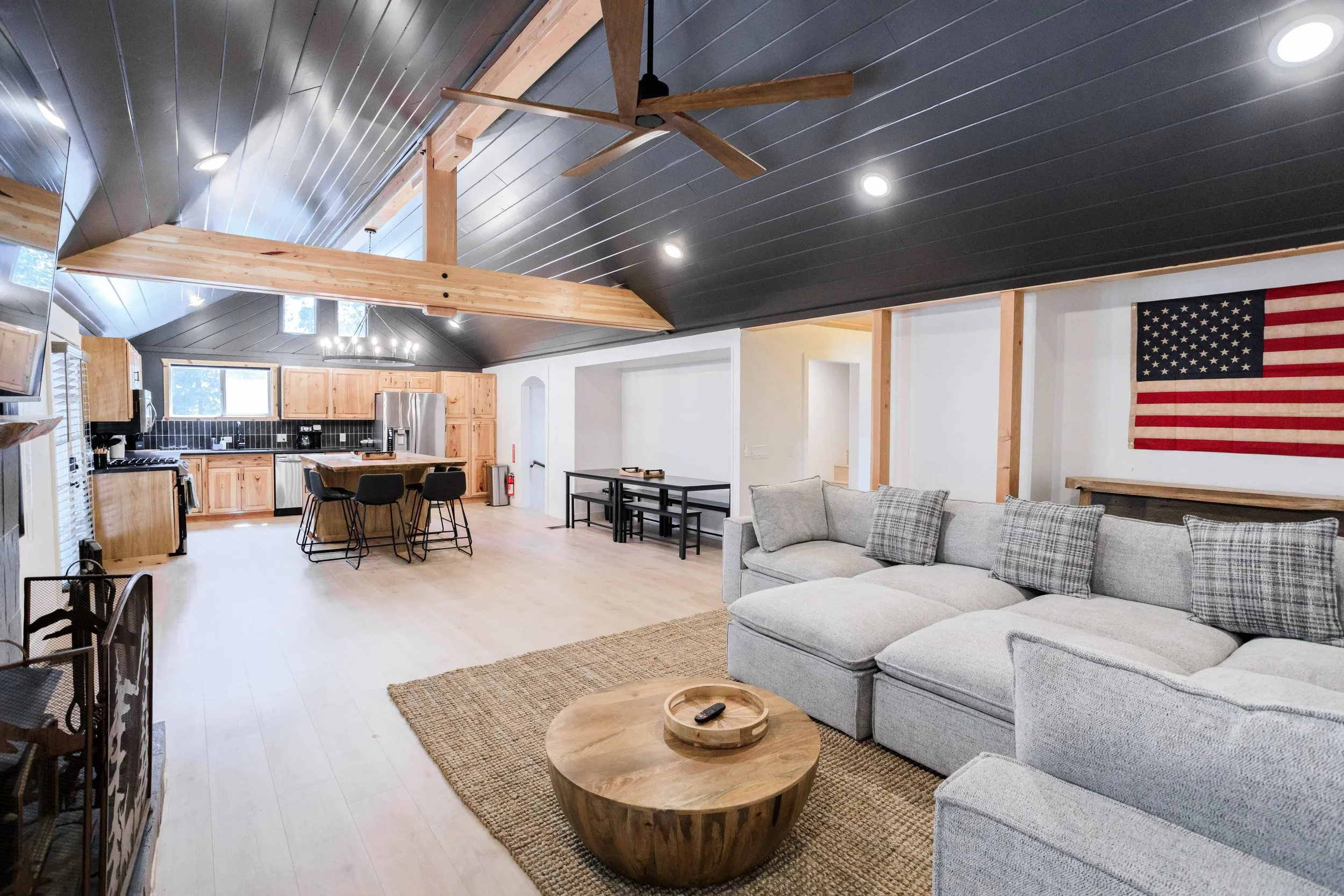 Open-concept living room and kitchen with black ceiling, wooden accents, American flag wall hanging, and light-colored furniture.