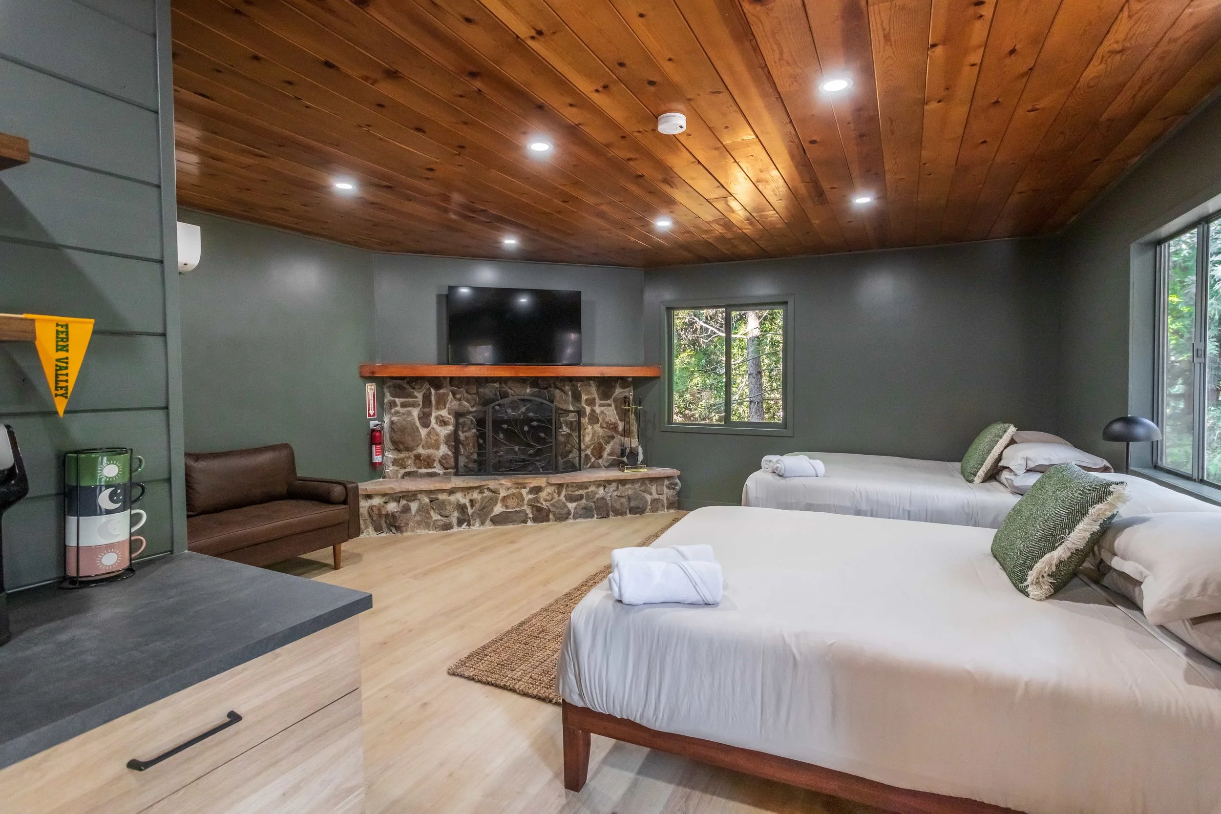 A cozy cabin room with two beds, a stone fireplace, a mounted TV, large windows showing trees outside, a small brown sofa, and a wooden ceiling.